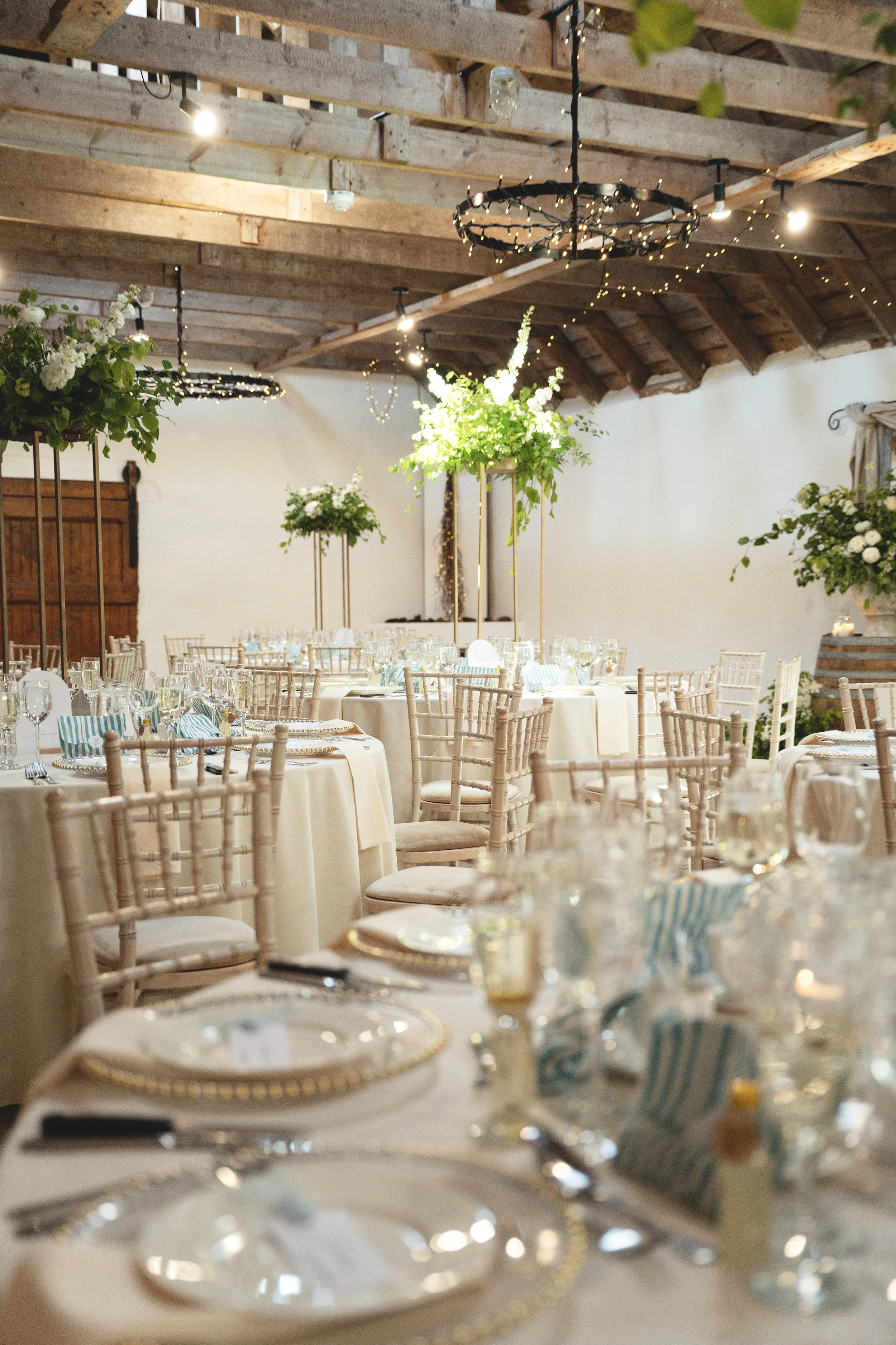 Elegant banquet hall decorated with large floral centerpieces on tall gold stands, round tables with cream tablecloths, and place settings with gold-rimmed chargers, glassware, and napkins. The ceiling has exposed wooden beams with string lights and 