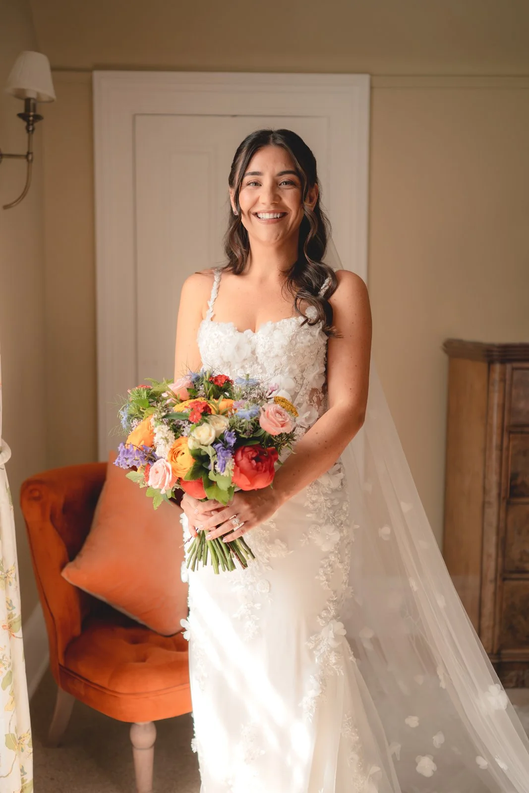 A smiling woman in a white wedding dress holding a bouquet of colorful flowers indoors.
