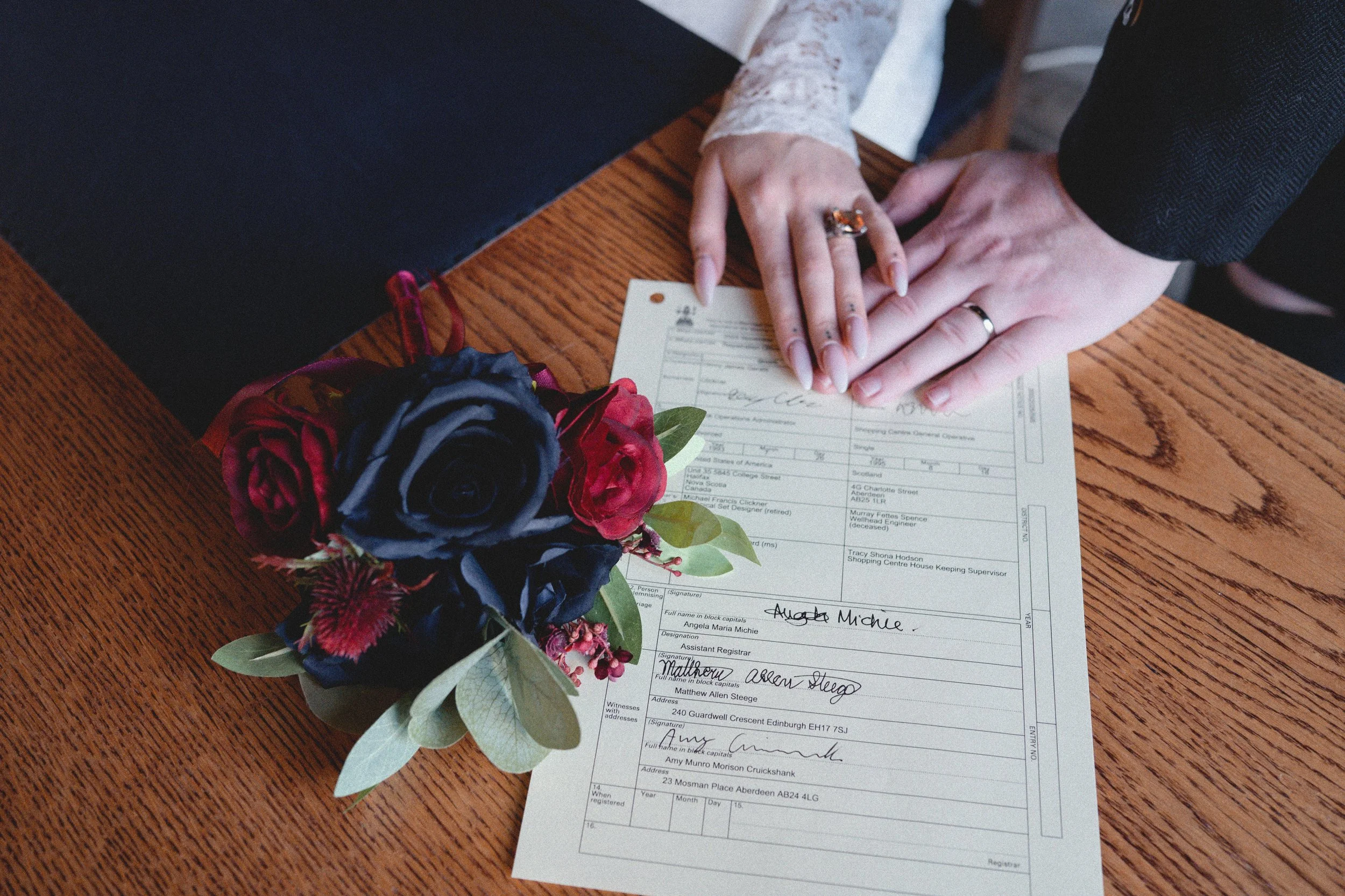 A couple's hands with wedding rings touching on a document, beside a bouquet of red and navy roses on a wooden table.
