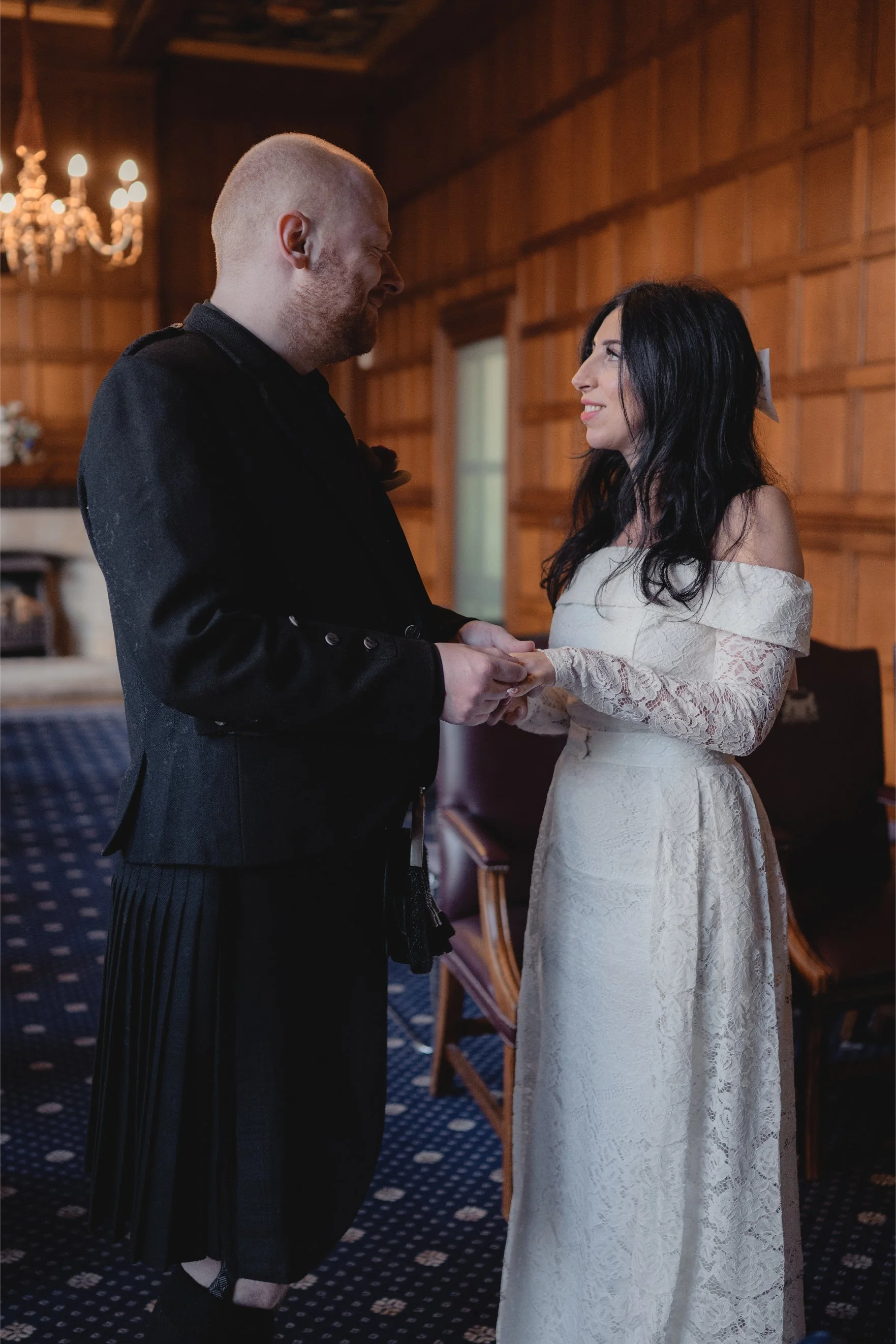 A man and woman standing inside a wood-paneled room, holding hands and smiling at each other, dressed in formal attire for a wedding or special occasion.