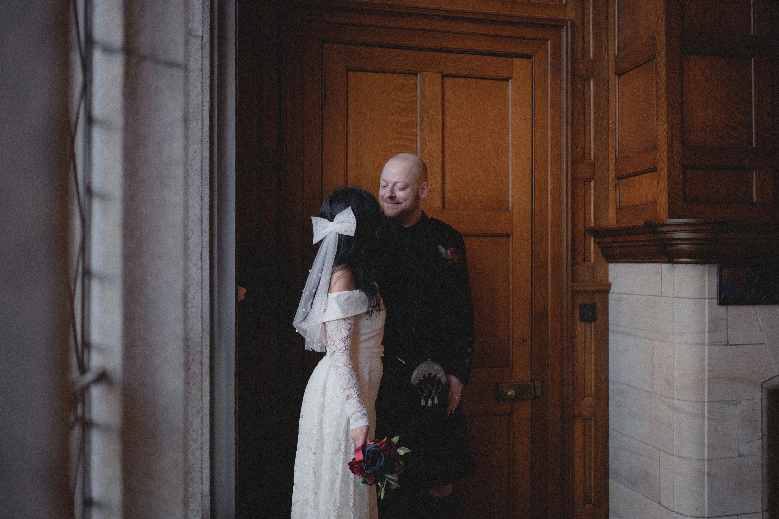 A bride and groom sharing a moment inside a wooden-paneled room. The bride is wearing a white lace wedding dress and holding a bouquet of dark red and purple flowers. The groom is dressed in a black kilt and shirt, smiling at the bride.