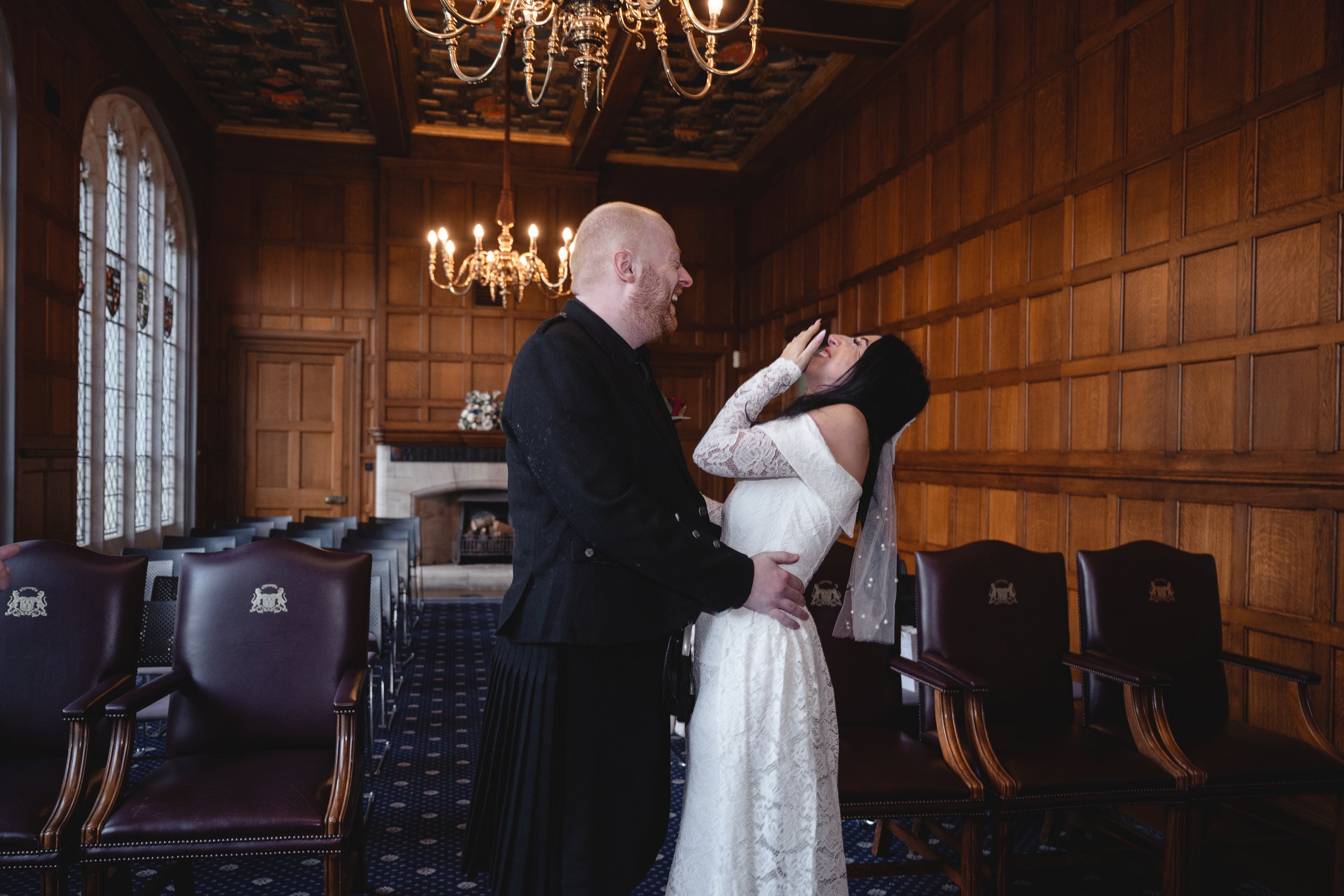 A bride and groom sharing a joyful moment in a wood-paneled room during their wedding ceremony. The bride is laughing and covering her mouth with her hand, while the groom smiles and holds her waist. The room has chandeliers, stained glass windows, a