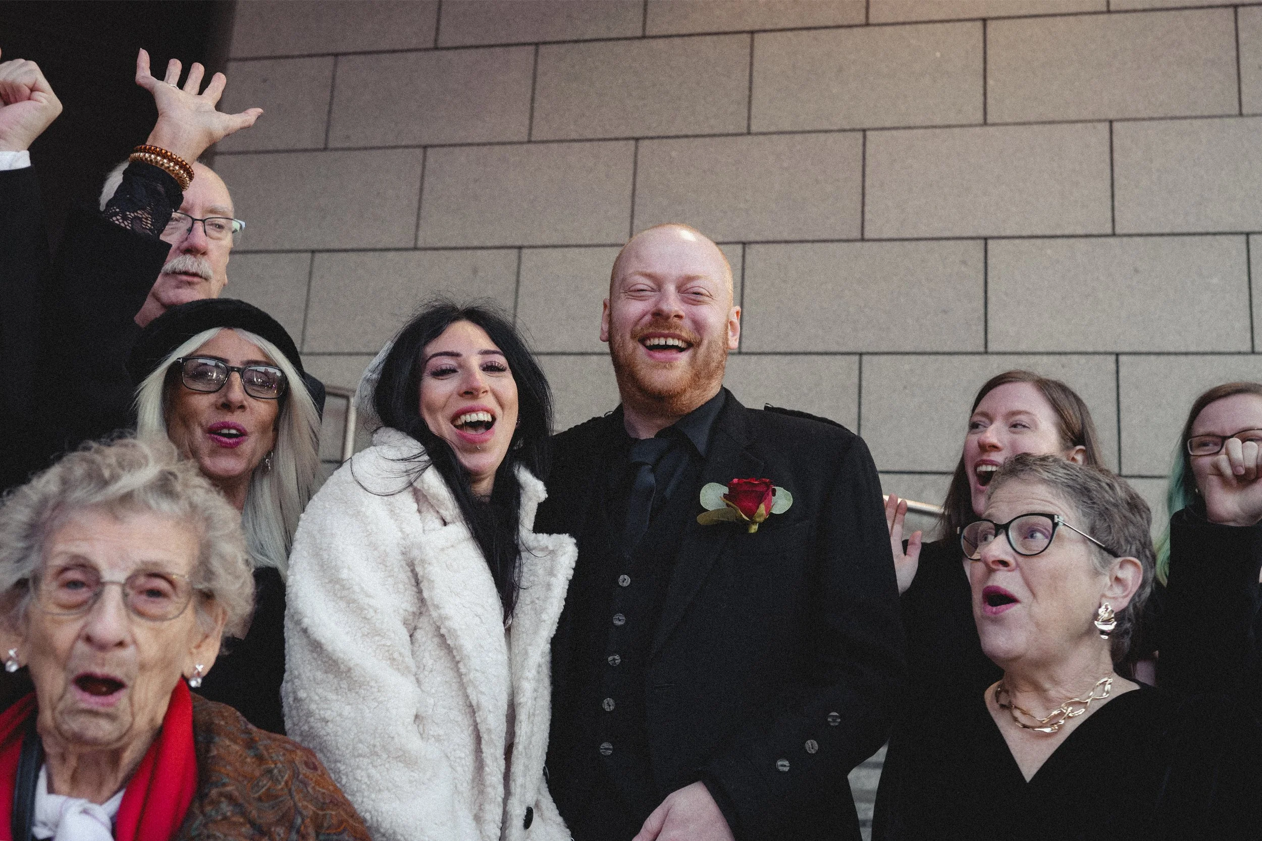 A group of people celebrating a wedding indoors. The bride and groom are smiling in the center, surrounded by family and friends.