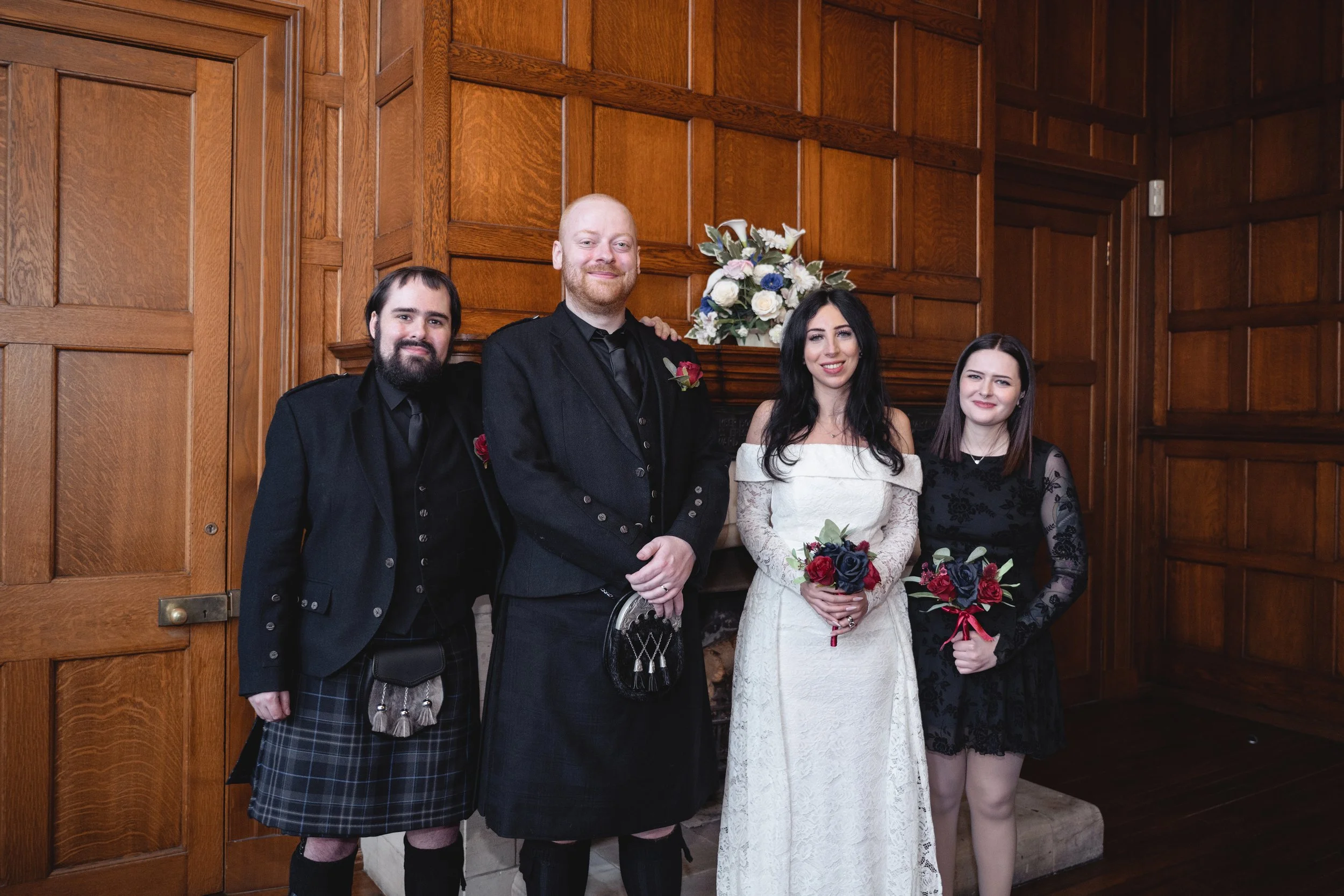 A group of four people dressed in formal attire standing in front of a wooden-paneled wall with a fireplace and a floral arrangement on the mantle. The group includes a man and woman in the center, possibly at a wedding or special event.