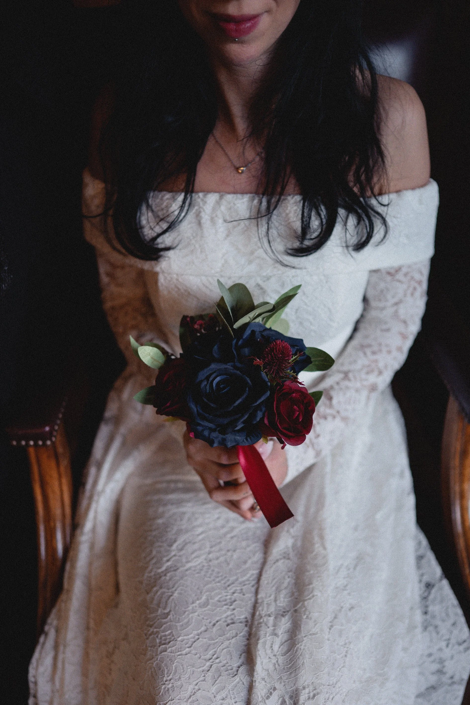 Woman in a white lace off-shoulder dress holding a bouquet of dark red and navy blue roses with greenery.