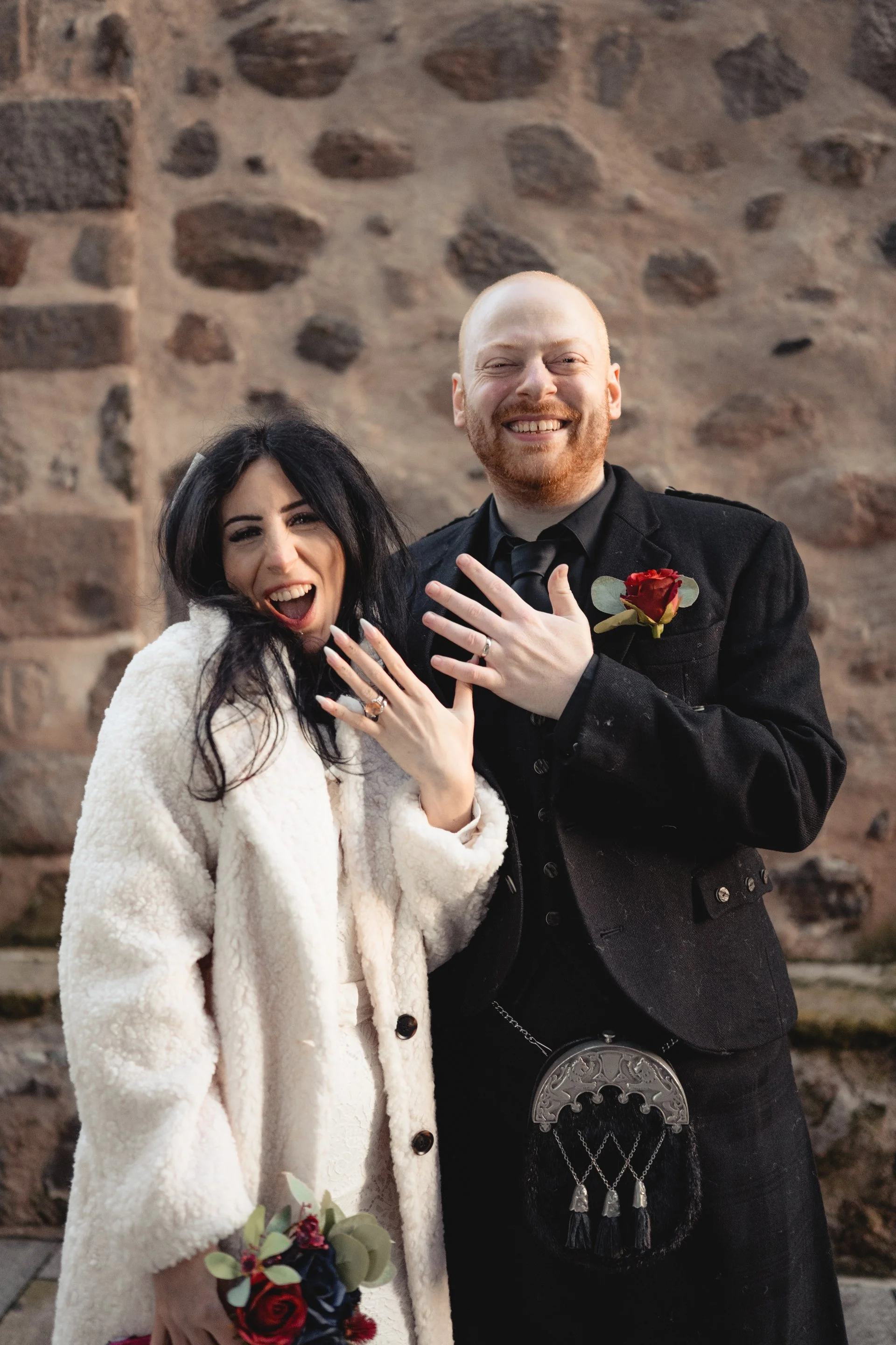 A happy couple celebrating their wedding, showing off their wedding rings, standing in front of a stone wall.