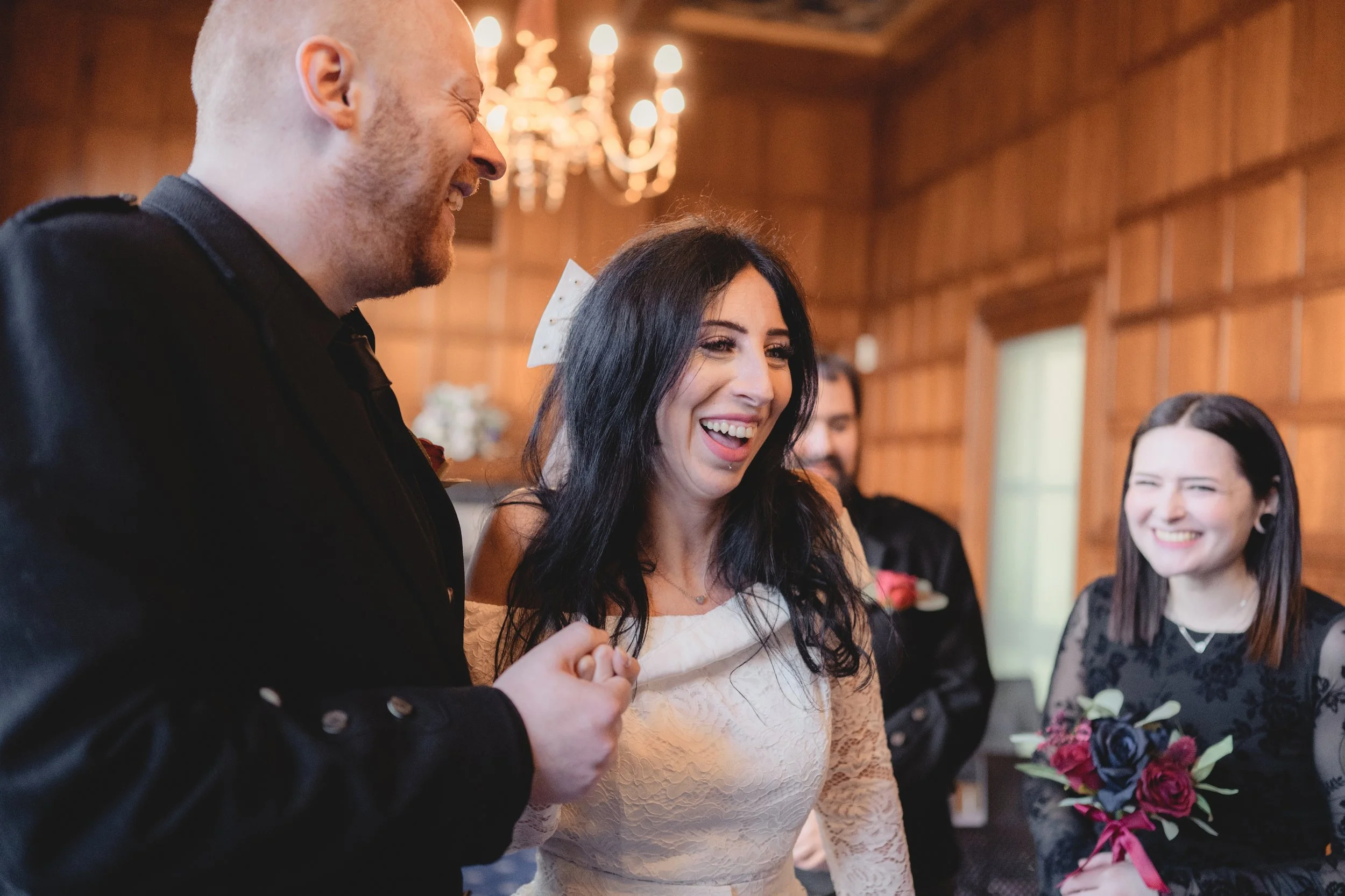 People smiling and enjoying a celebration indoors, with a woman in a white dress and others in black dresses, in a warmly lit room with wood-paneled walls.
