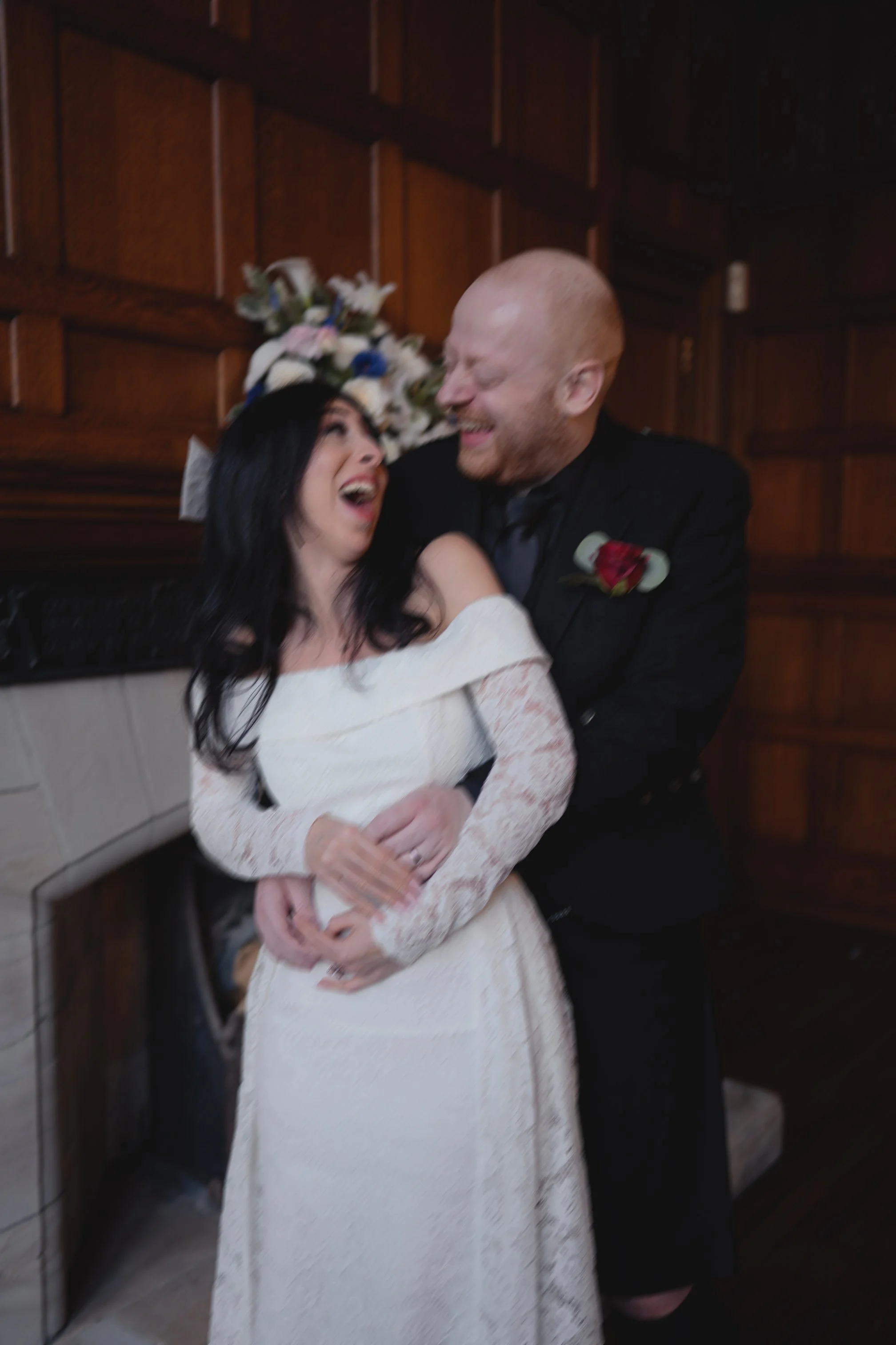 Happy couple at wedding, the woman in a white lace dress and the man in a black tuxedo, embracing and laughing in front of a wooden wall and floral arrangement.