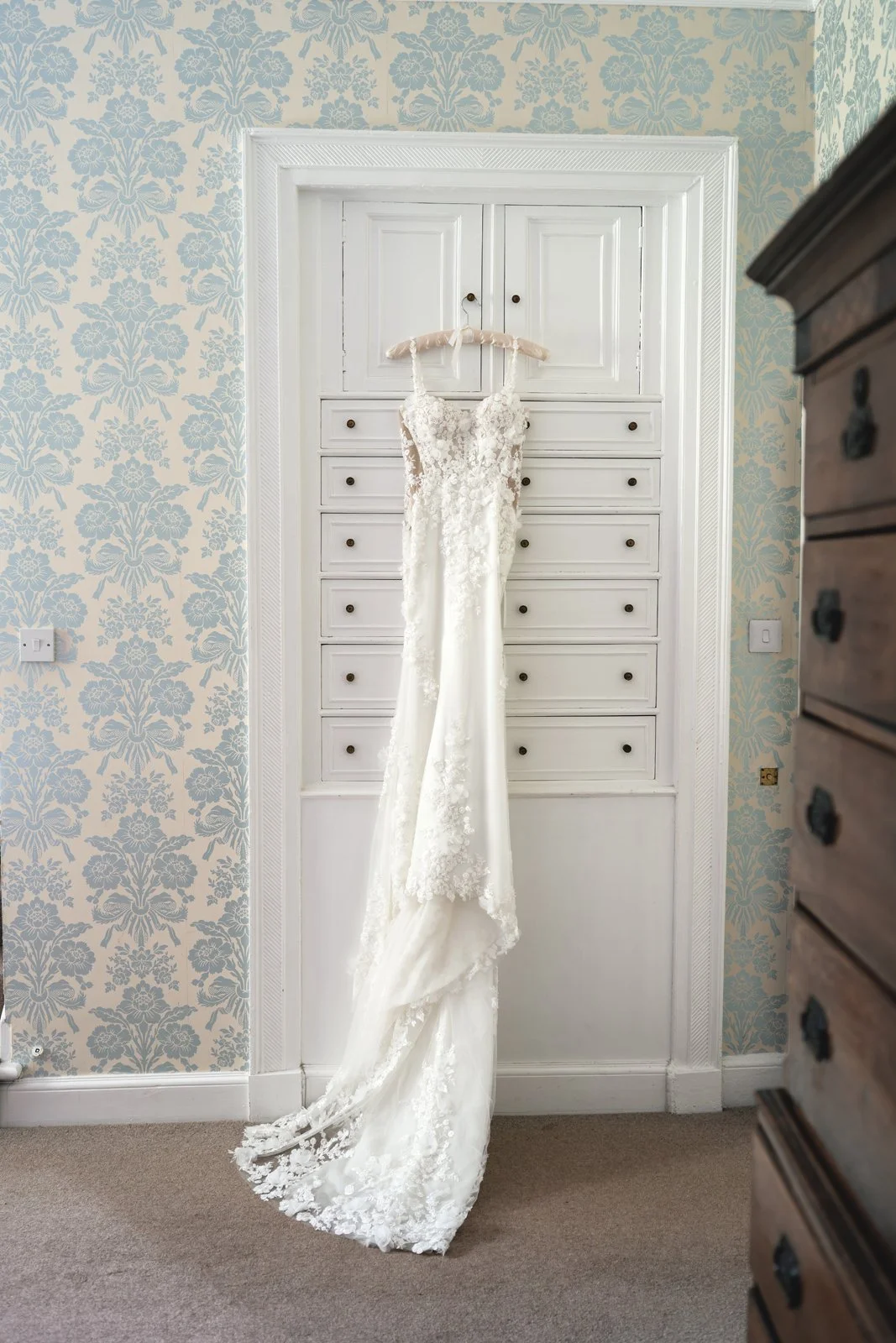 A white wedding dress hangs on a padded hanger from a white dresser in a room with light floral wallpaper.