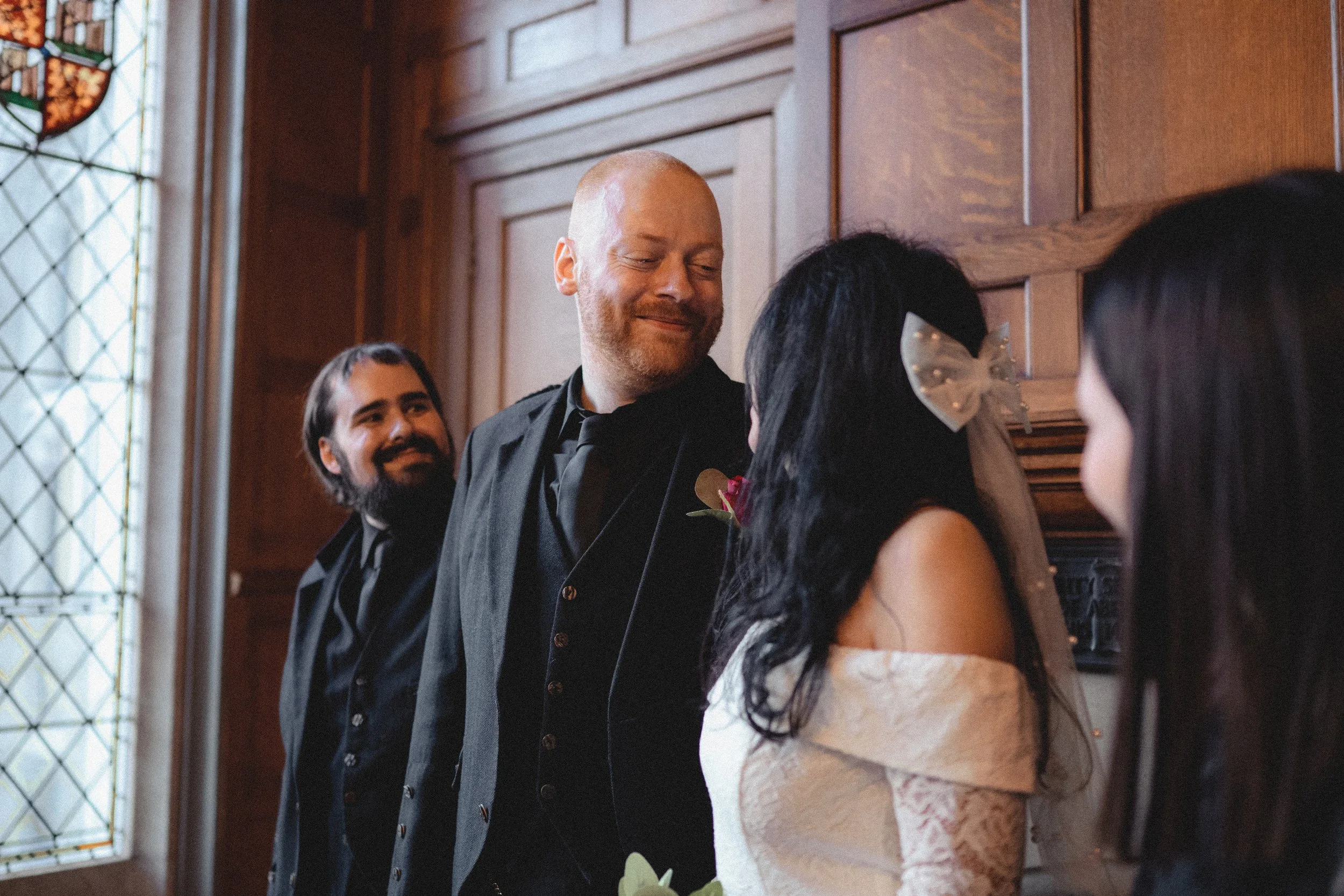 A wedding ceremony with a couple at the altar. The groom is smiling, balding with a beard, wearing a black suit. The bride is wearing a white off-shoulder dress with a veil and bow, her face is not visible. Two women stand beside them, one with dark 