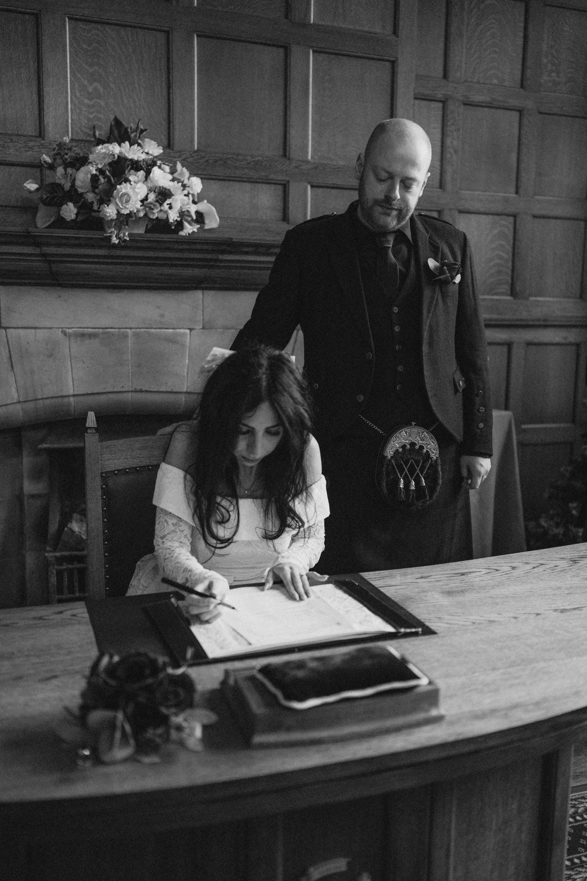 A woman sitting at a desk signing a document while a man dressed in formal attire and a kilt stands beside her, in a wood-paneled room with a fireplace and a floral arrangement.
