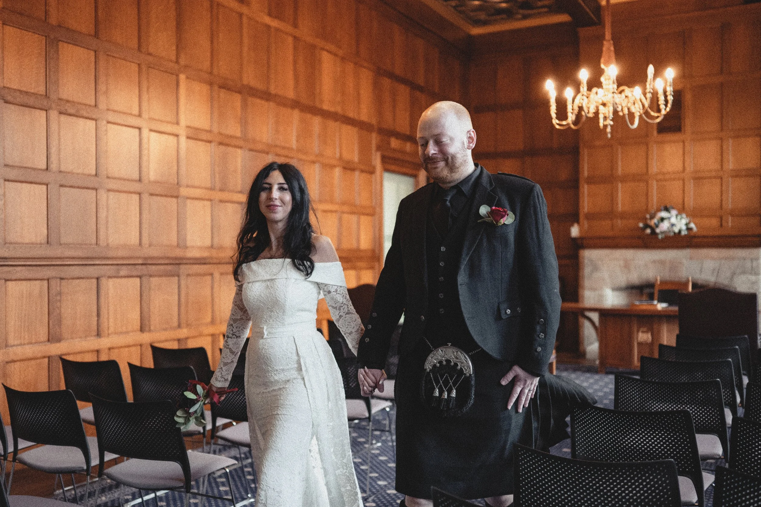 A bride in an off-the-shoulder white lace wedding dress and a groom in a black Scottish kilt and jacket, holding hands, standing in a wood-paneled room with a chandelier hanging from the ceiling and empty chairs arranged in rows.