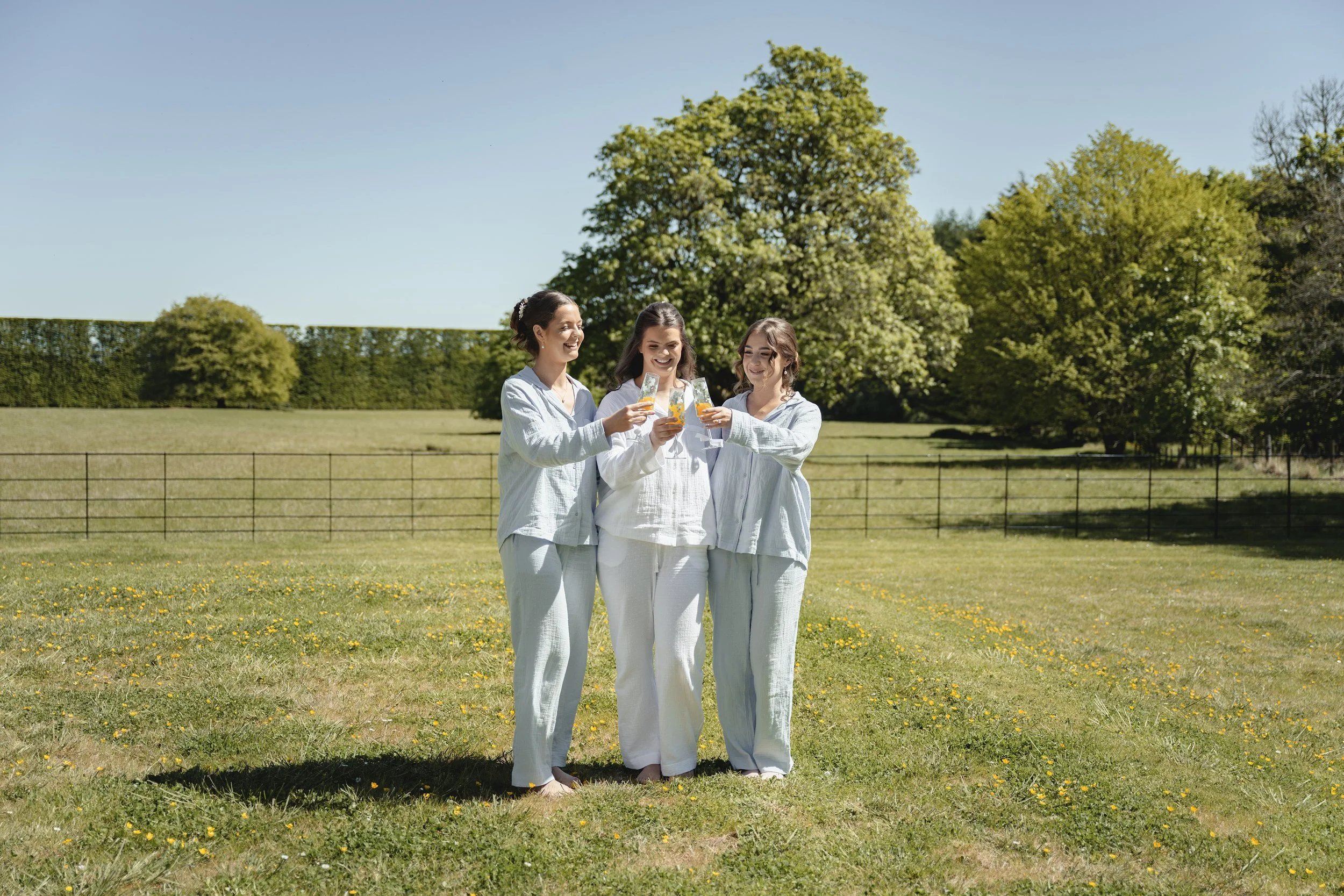 Three women in pajamas standing on a grassy field holding glasses of beverages, smiling and toasting.