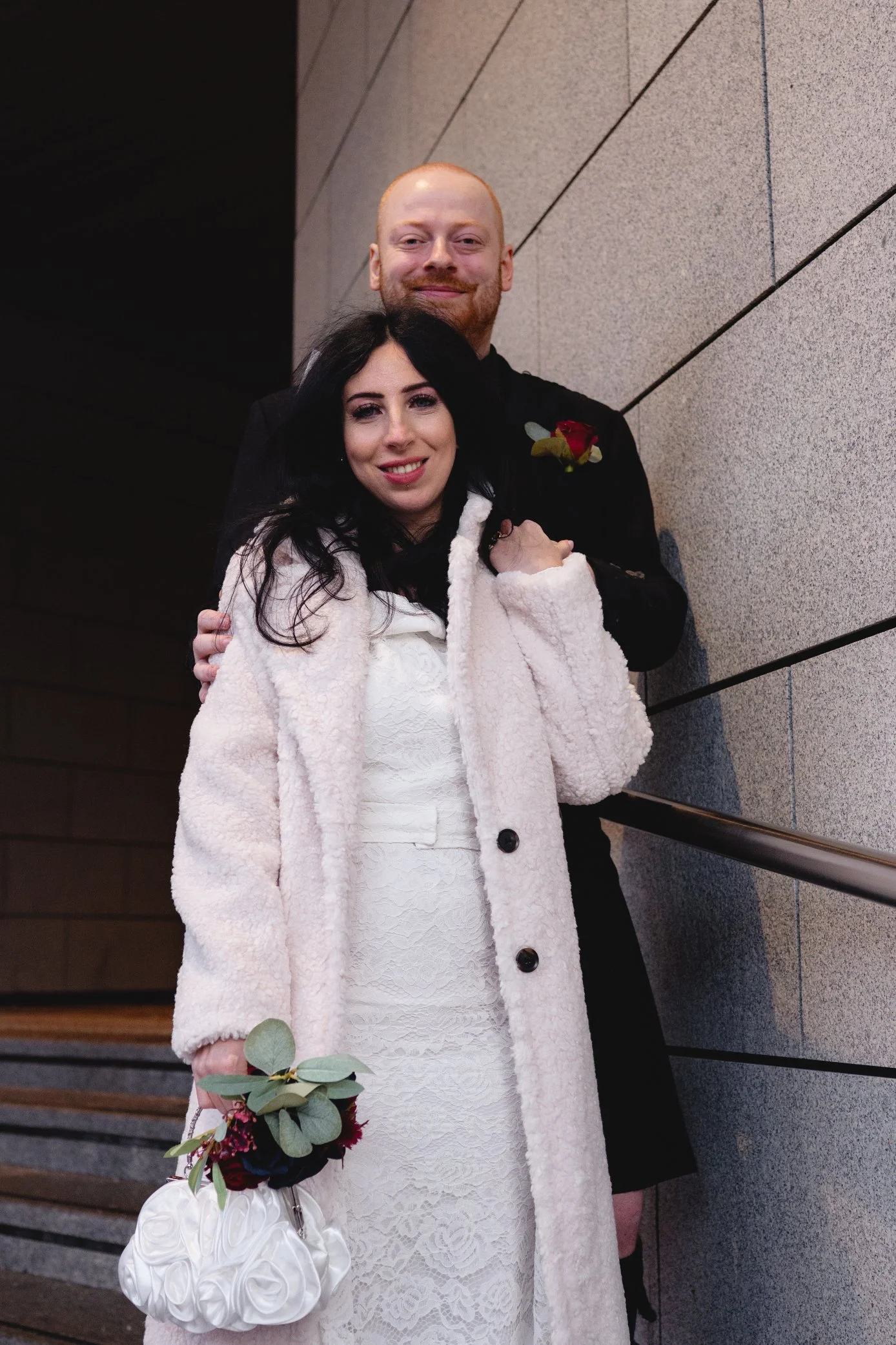 A man and woman standing on stairs, smiling at the camera. The woman is holding a bouquet and wearing a white lace dress with a cream-colored coat. The man is dressed in black with a boutonniere on his lapel.
