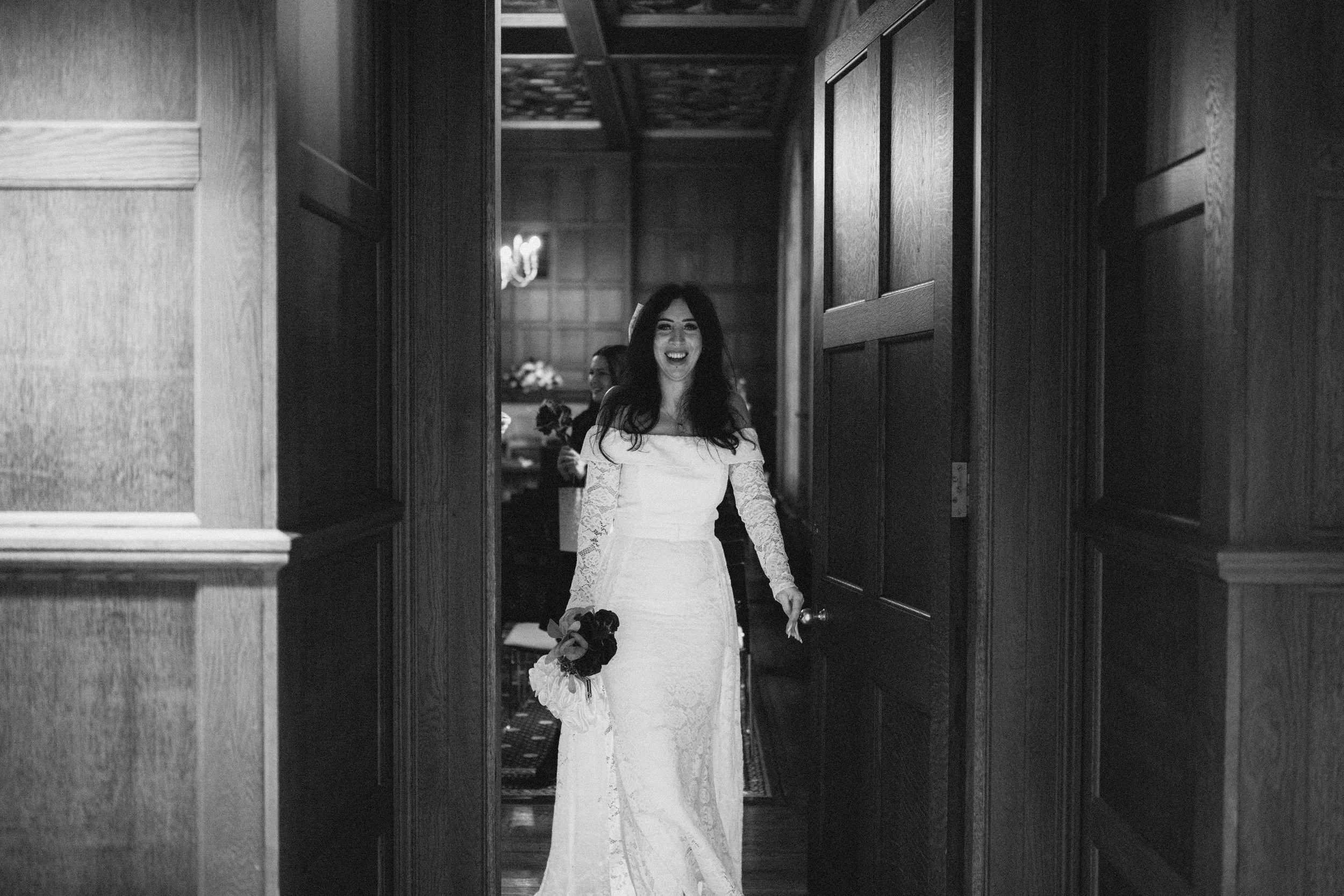 A woman in a white wedding dress holding a bouquet, smiling as she enters a room.