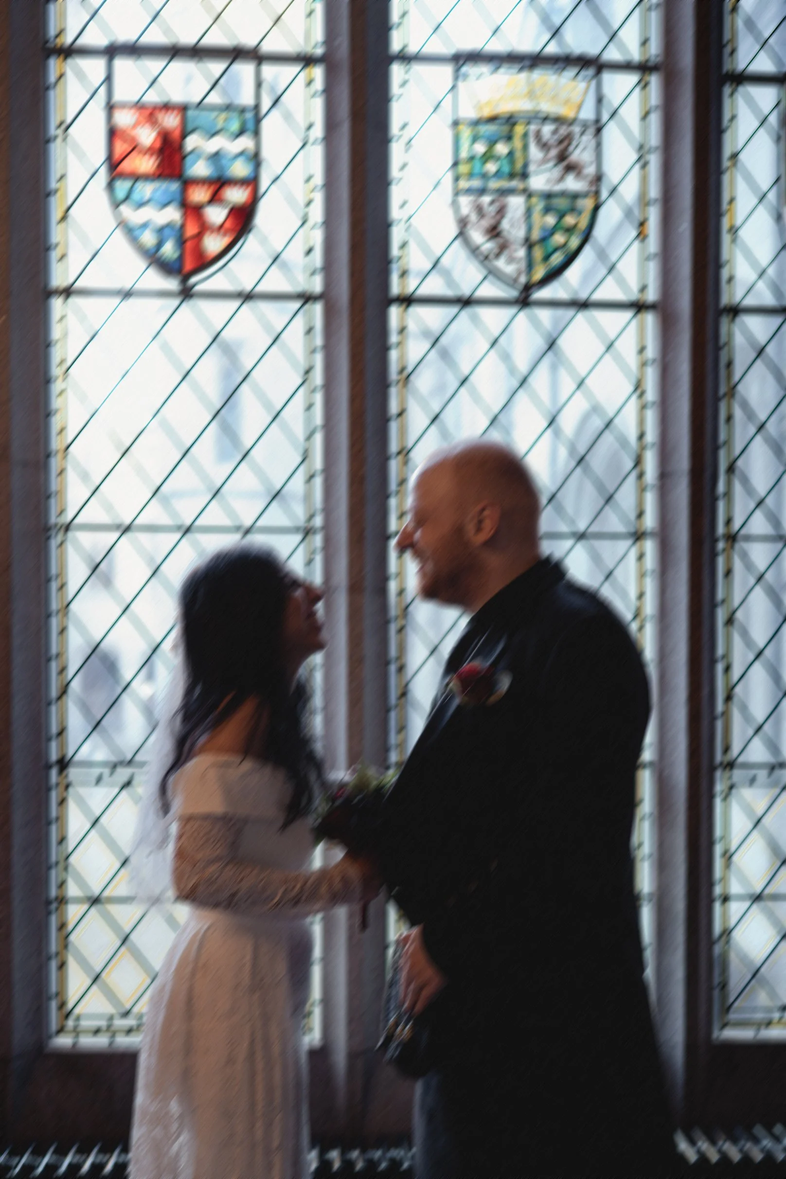 A couple in wedding attire holding hands inside a church with stained glass windows featuring heraldic shields.