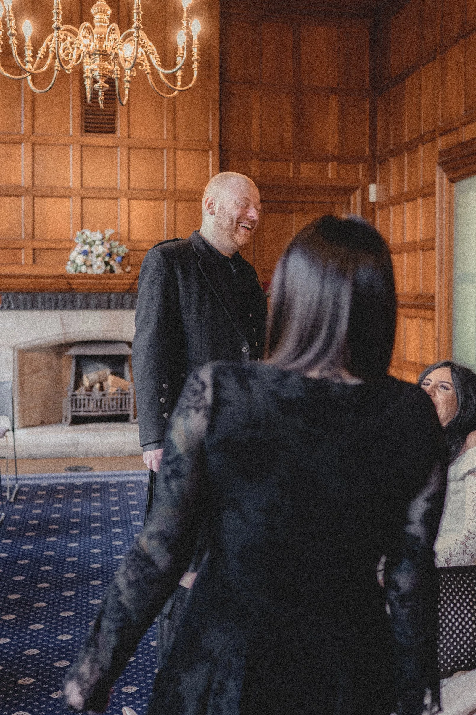 A man with a beard and bald head is smiling and laughing, in a black blazer, standing in a wood-paneled room with a chandelier and fireplace, as two women face him, one with dark hair and black lace clothing, the other partially visible with dark hai