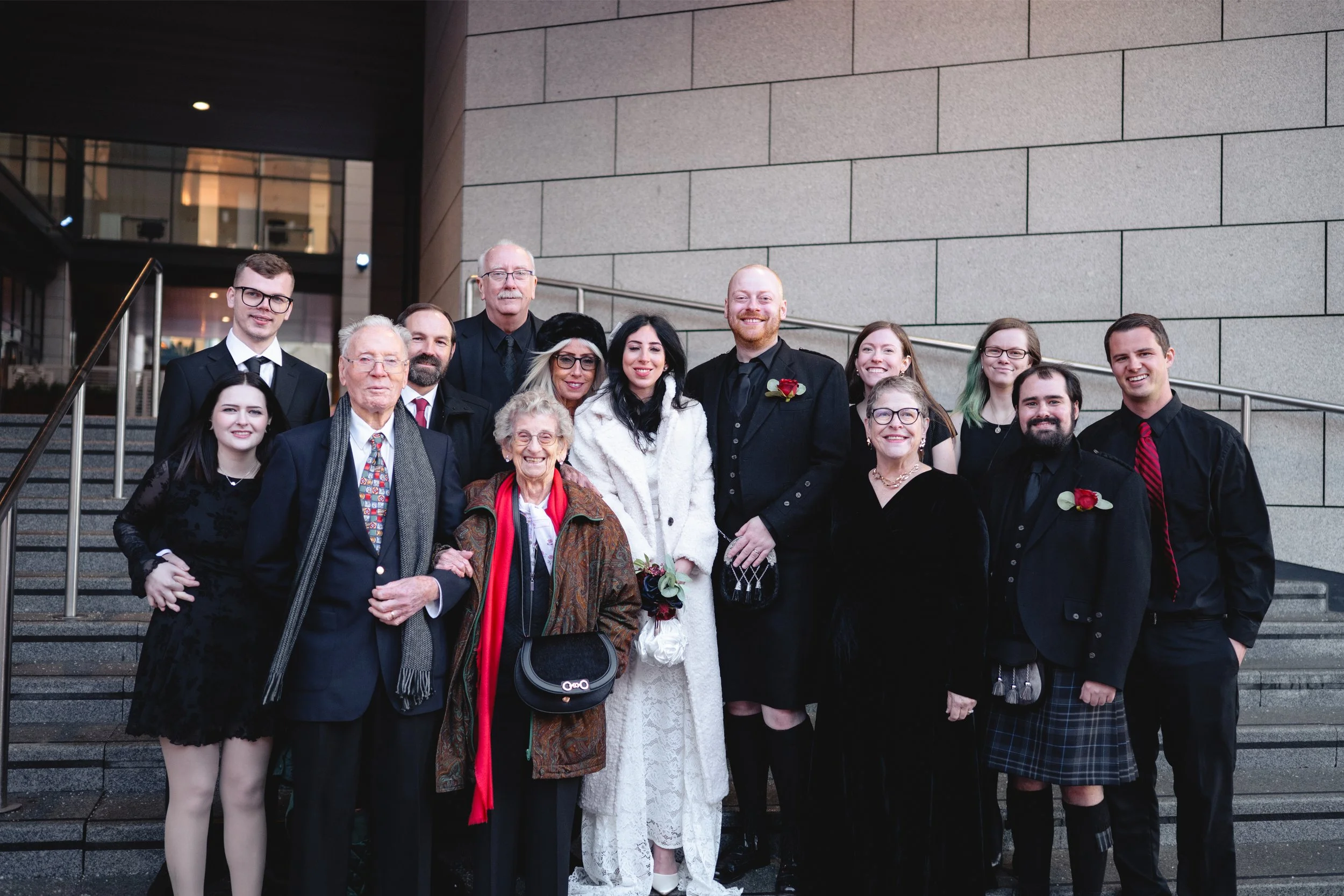 A group of people celebrating a wedding on the steps of a building. The bride and groom are in the center, with the bride wearing a white dress and coat, and the groom in a black suit with a red boutonniere. The group includes men and women dressed i