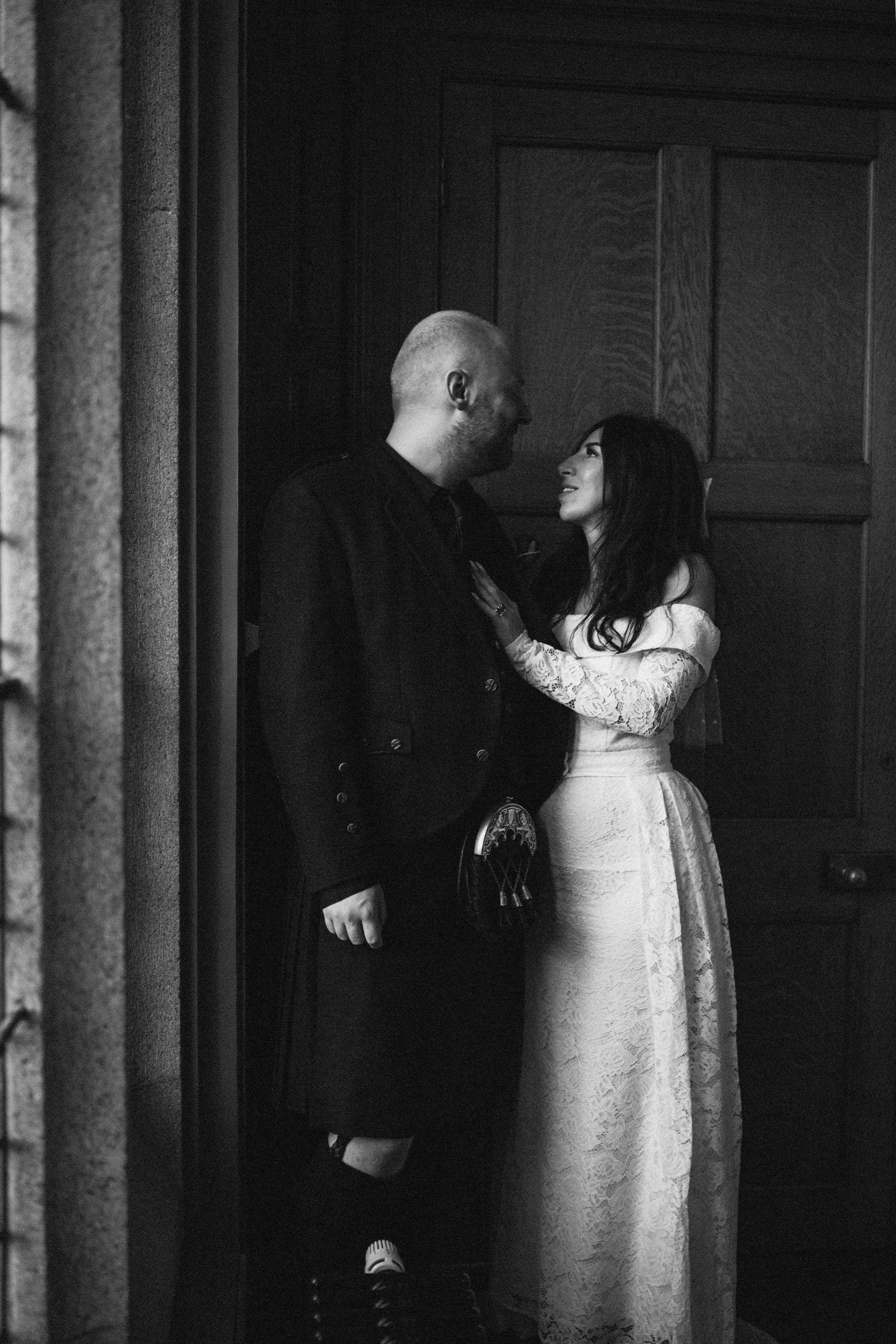 A black and white photo of a couple in vintage attire standing close and looking at each other affectionately inside a room with wooden paneling.