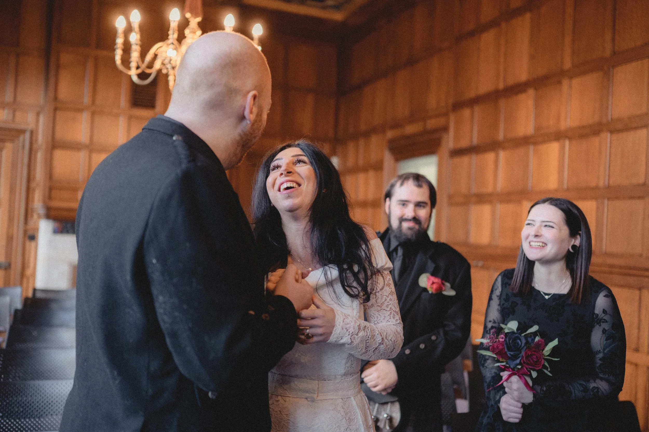 A couple exchanging vows at a wedding ceremony, with two other guests smiling in a wood-paneled room.