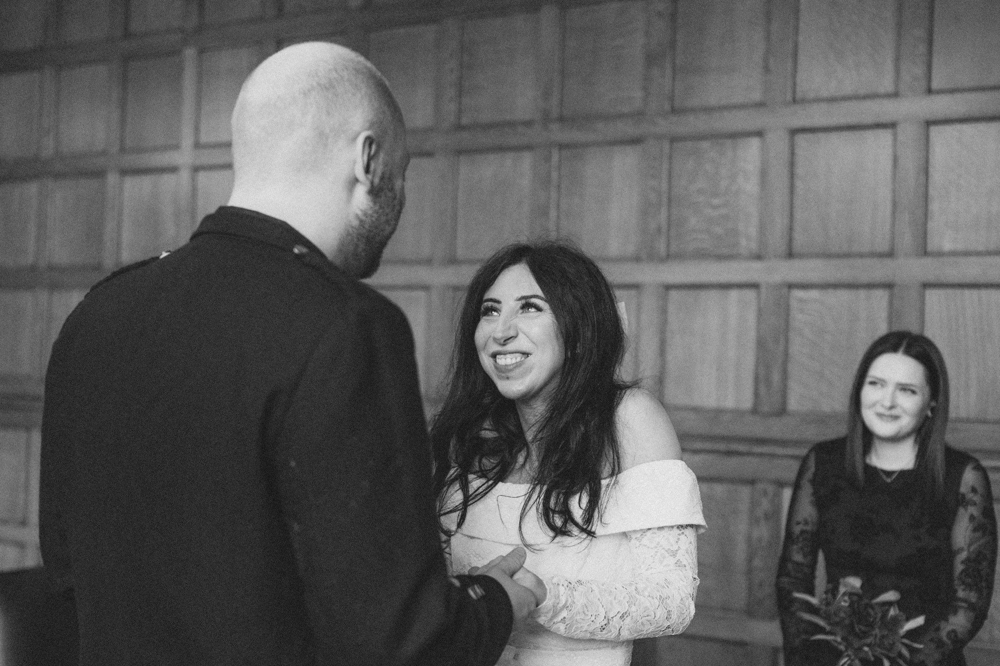 A woman in a white off-shoulder lace dress is smiling and looking at a man with a closely shaved head, dressed in black, who is facing her. In the background, another woman with long dark hair, wearing a black lace dress, is watching them and smiling