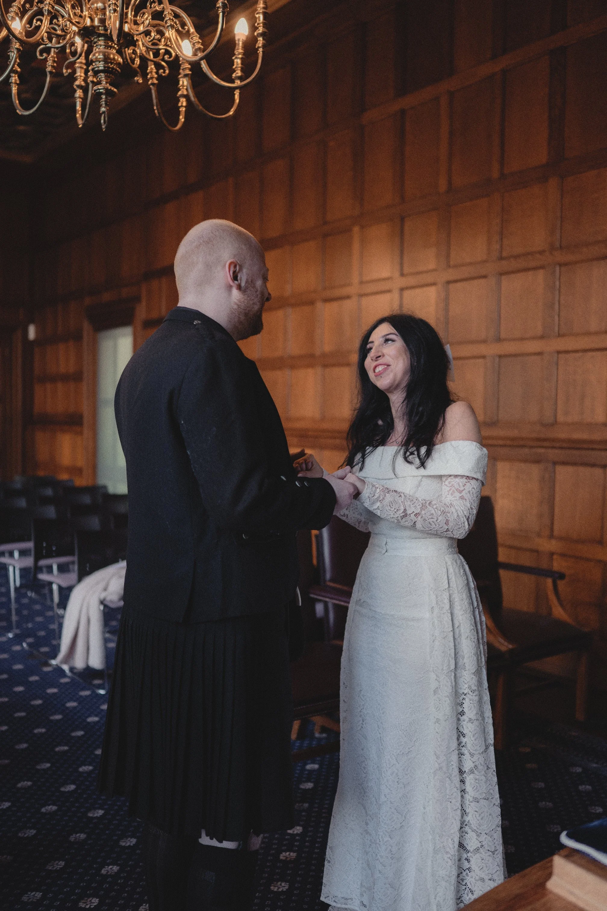 A bride and groom stand together in a wood-paneled room, holding hands and gazing at each other during their wedding ceremony.