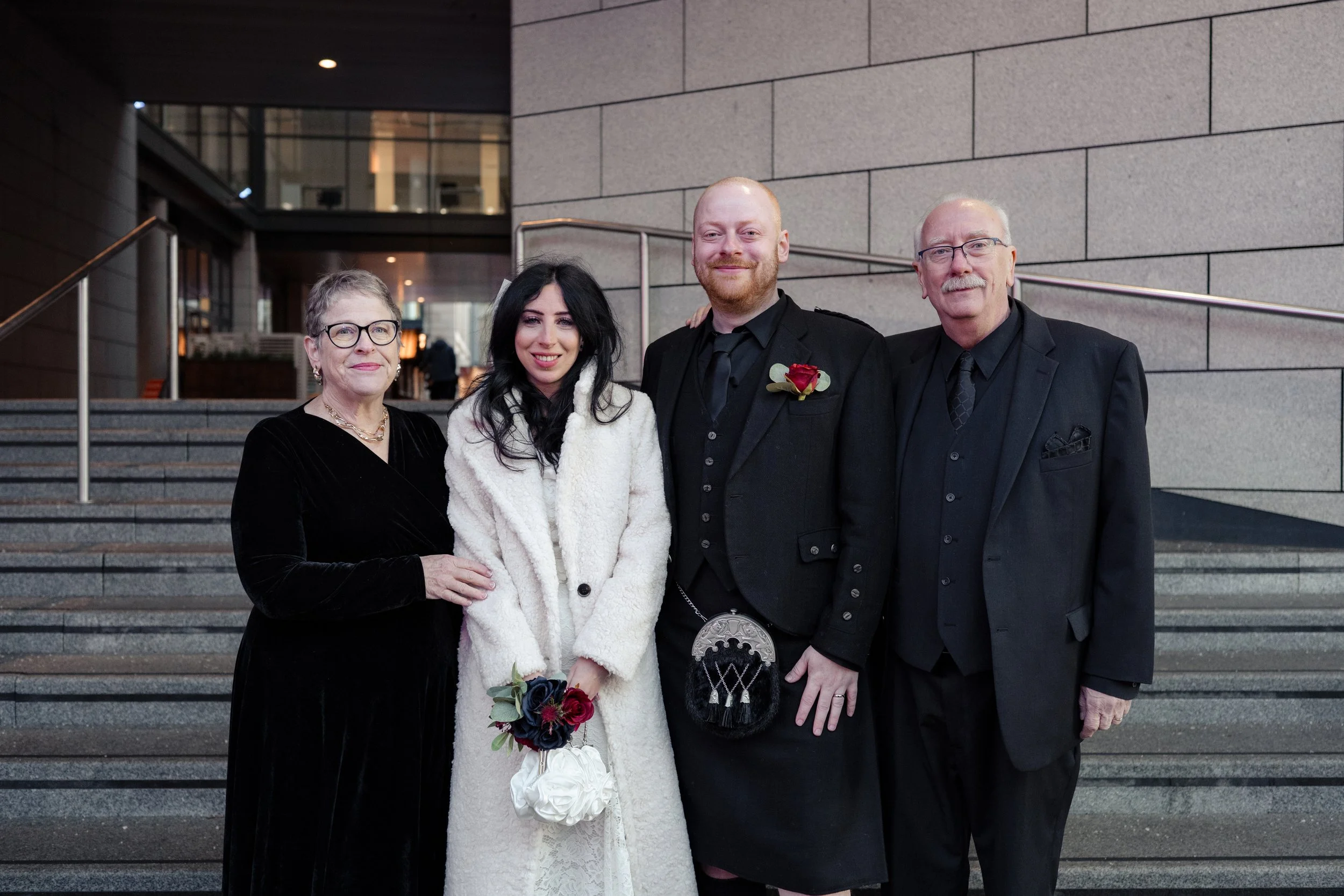 Four people in formal attire posing for a photo on a modern building stairway with glass and concrete walls in the background.