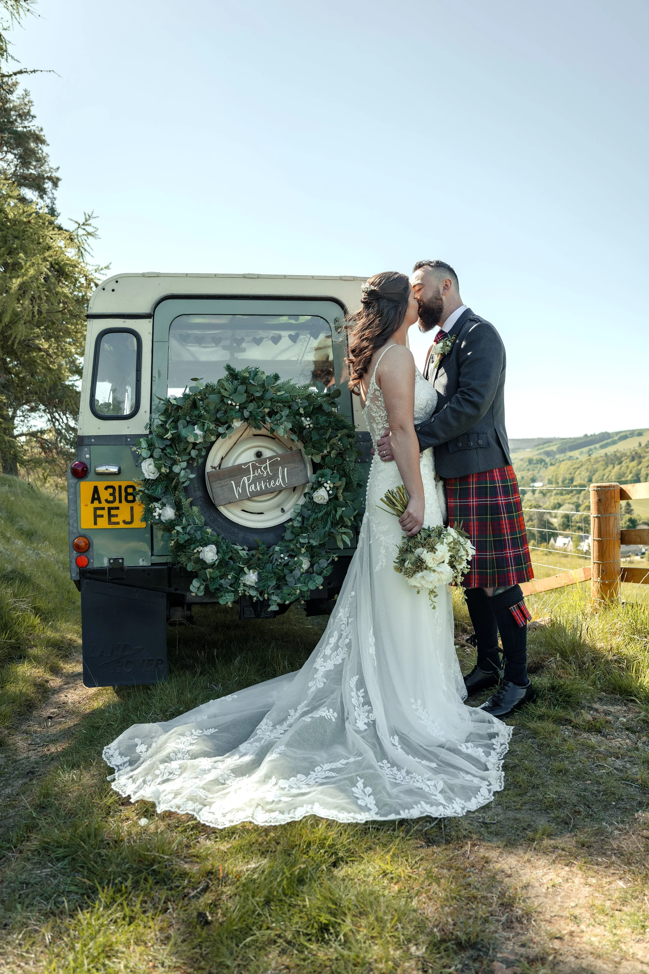 A bride and groom kissing in front of a vintage land rover decorated with green foliage and white roses, outdoors on a bright, sunny day.