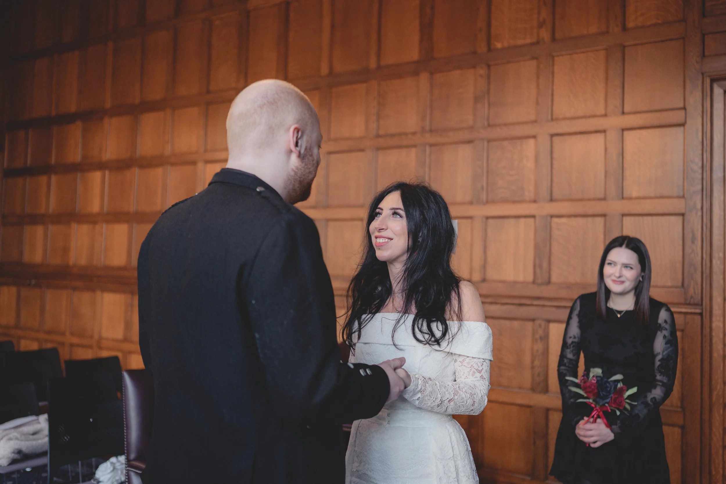 A bride and groom exchanging vows, with a woman holding a bouquet of roses in the background during a wedding ceremony in a wood-paneled room.