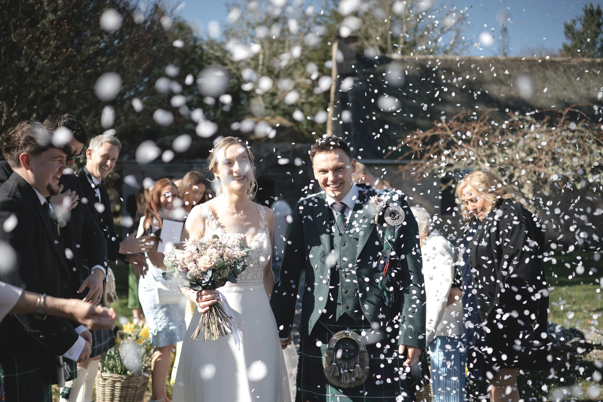 A newlywed couple celebrating outdoors with friends during their wedding, surrounded by falling confetti.