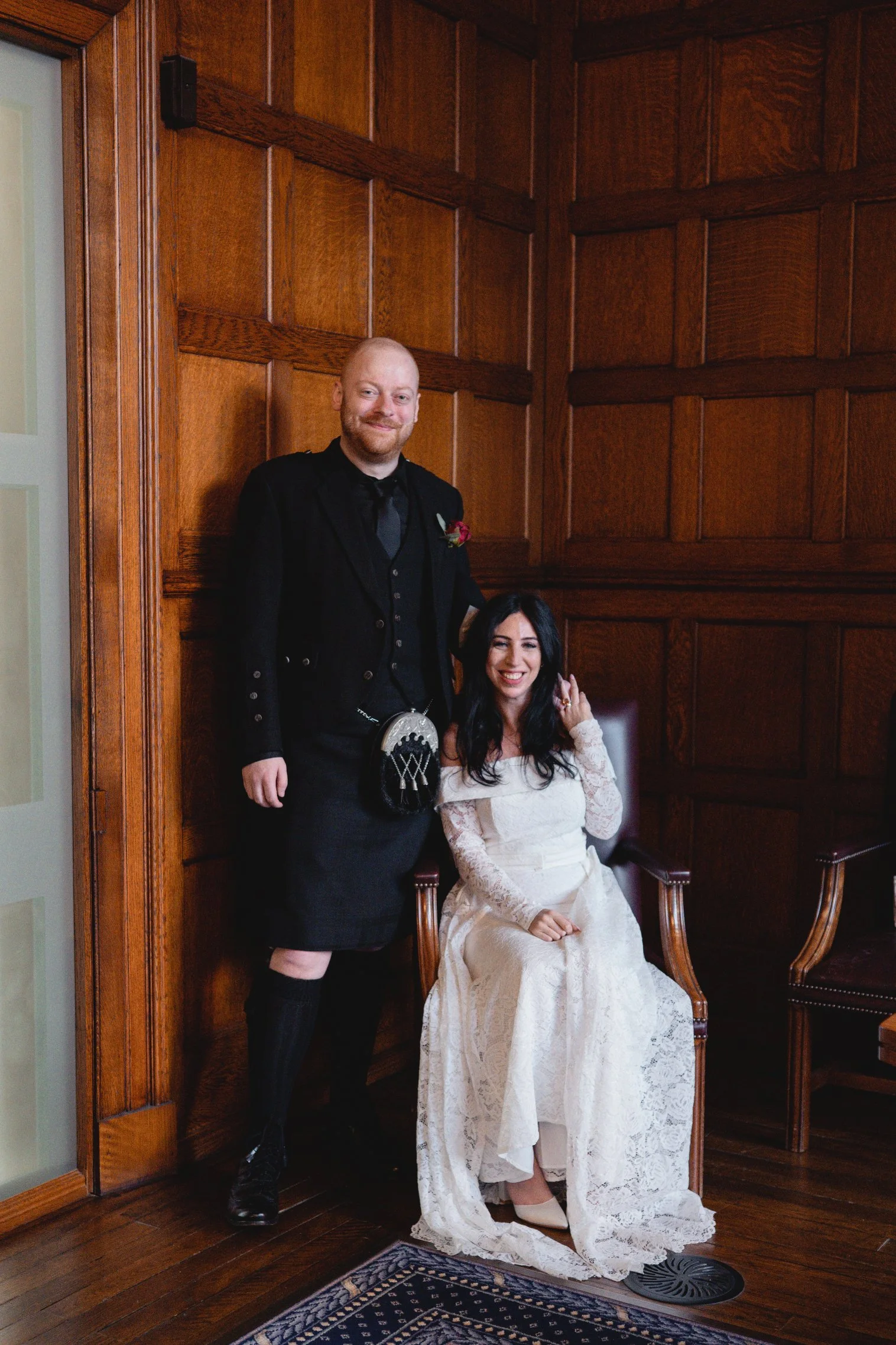 A man and woman in wedding attire inside a wood-paneled room. The woman is sitting in a chair, smiling, wearing a white lace wedding dress. The man stands beside her, dressed in a black jacket and kilt, smiling.