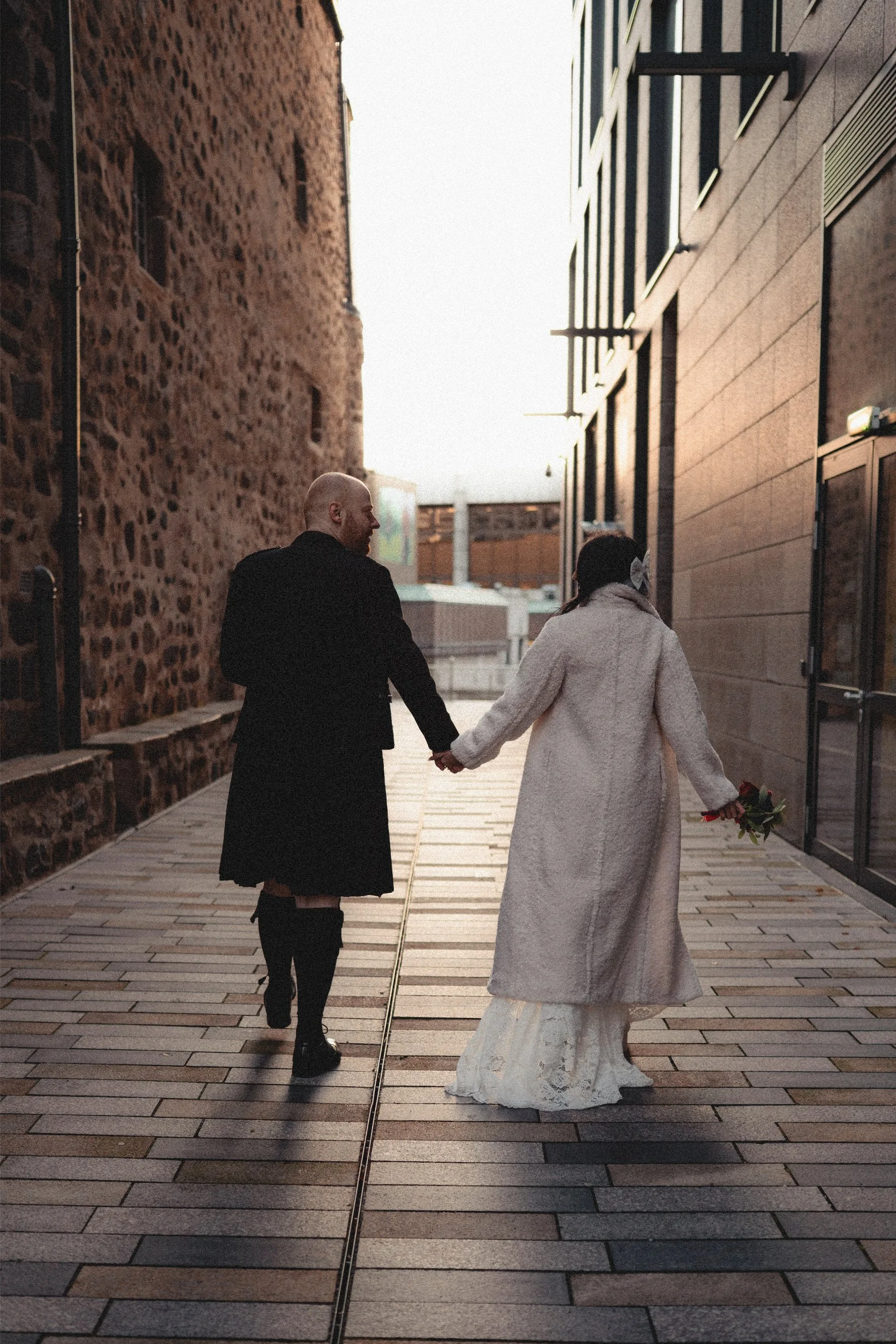 A couple holding hands and walking away on a city sidewalk during sunset. The woman is wearing a long white coat and a white dress, holding a bouquet of flowers. The man is dressed in a black coat and boots. They are walking between a stone building 