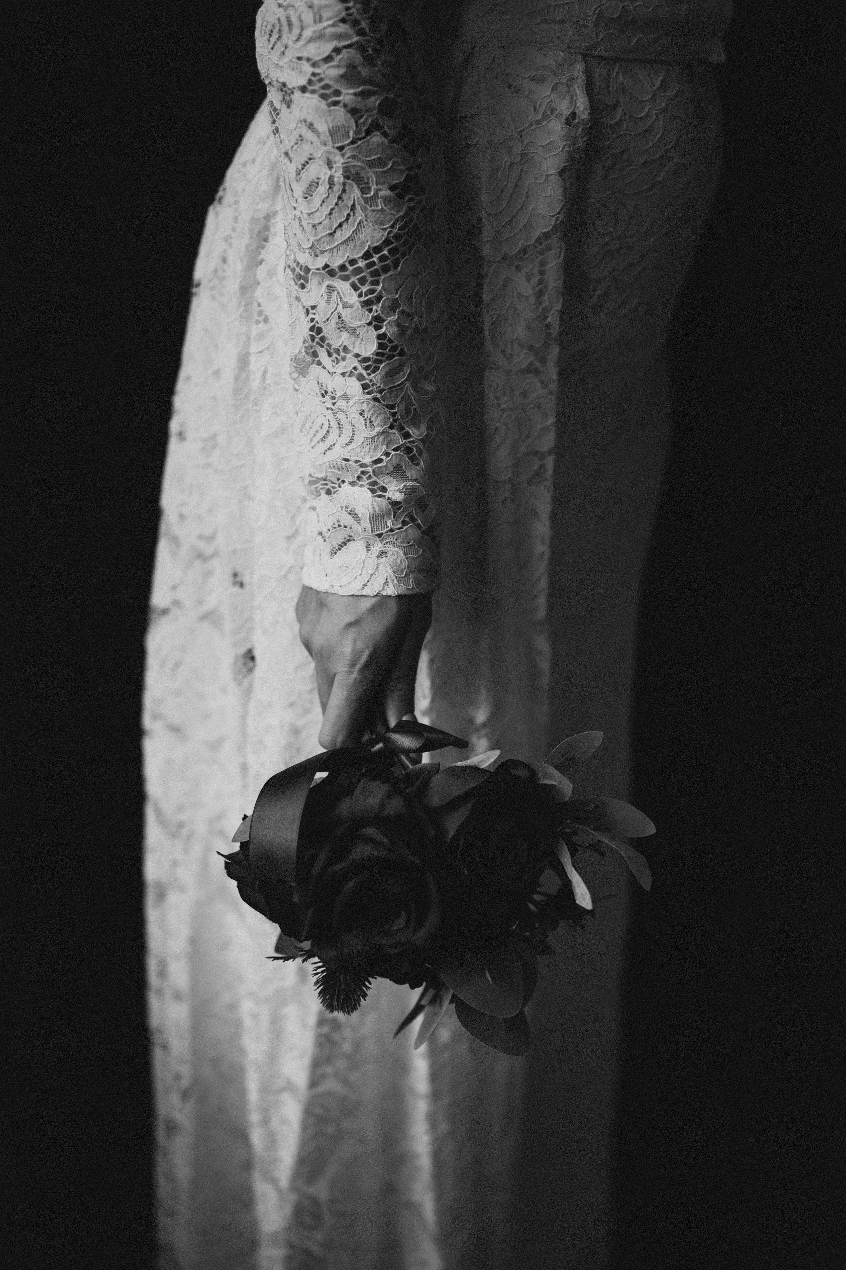 A person wearing a lace dress holding a small bouquet of dark flowers in their hand, against a dark background.