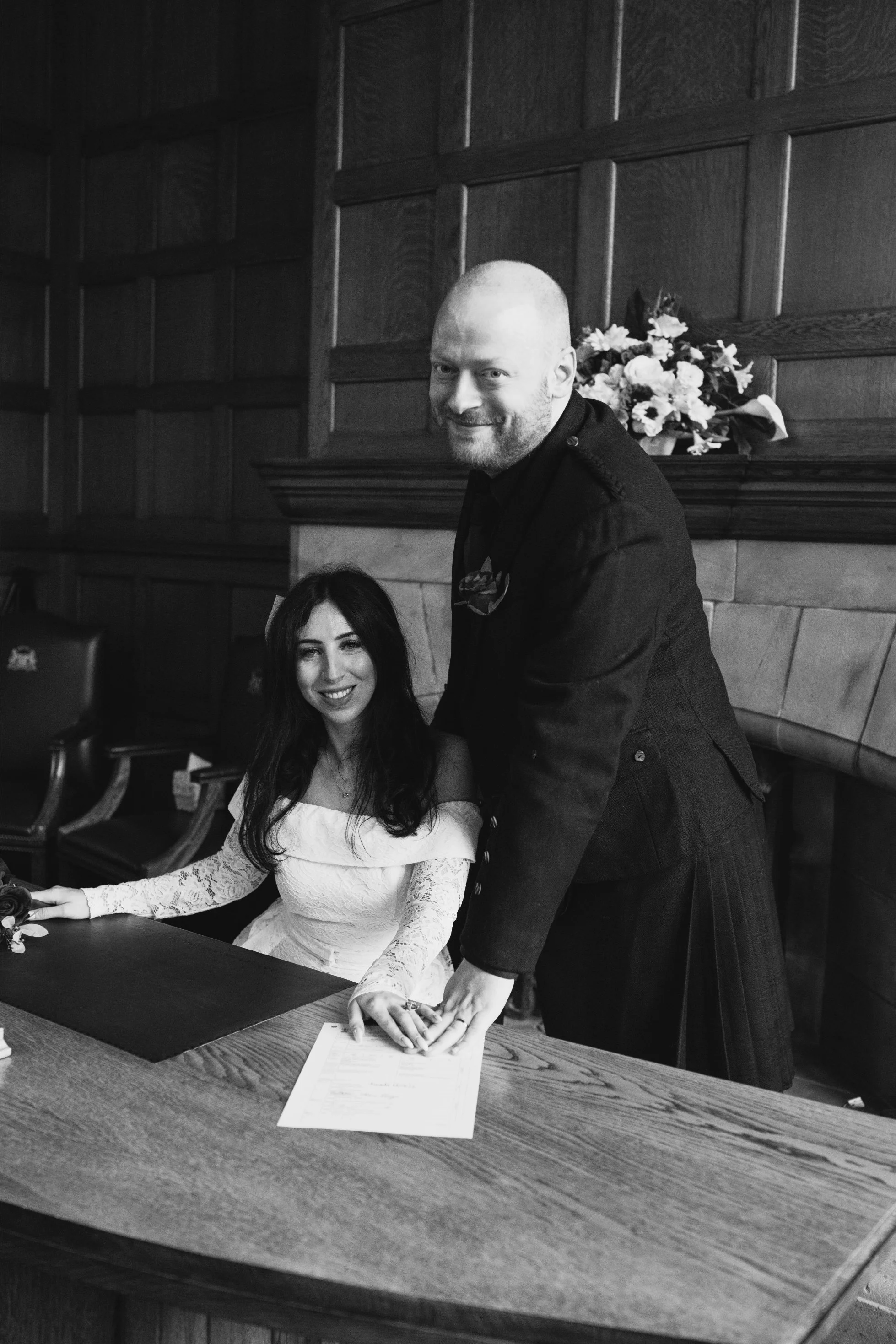A woman in a wedding dress sitting at a table with a man in a dark suit standing beside her, both smiling, in a room with wood-paneled walls and a fireplace with a flower arrangement above it.