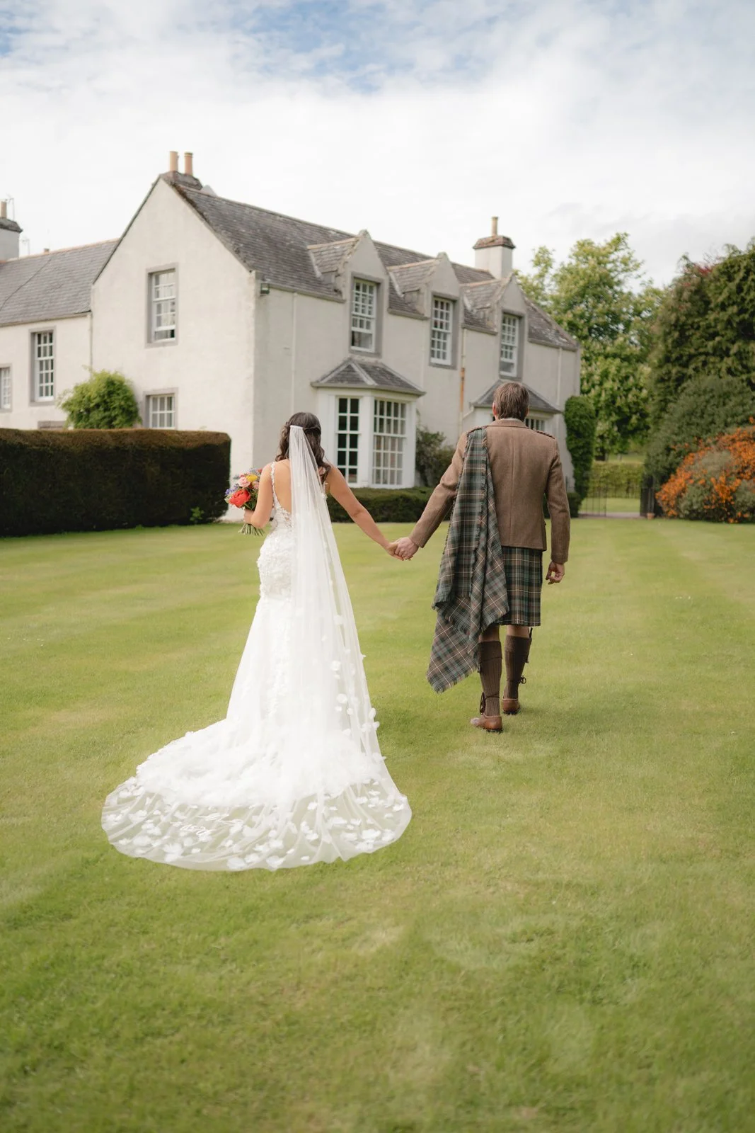A bride and groom walk hand in hand across a lawn outside a large white house, with the bride wearing a white wedding gown and veil holding a bouquet, and the groom in traditional Scottish attire with a tartan kilt and shawl.