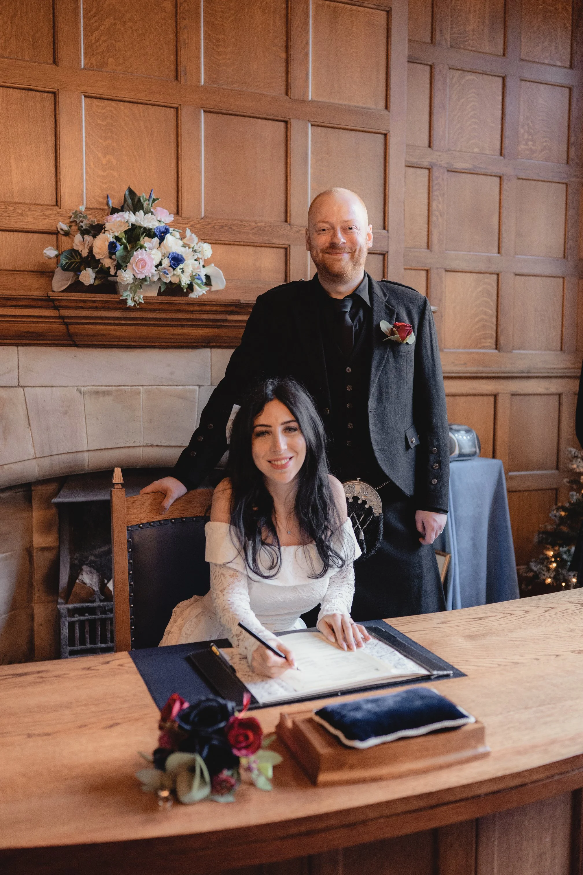 A woman in a white dress signs a document at a table, while a man in a black suit stands behind her. They are in a wood-paneled room with flowers and holiday decorations.