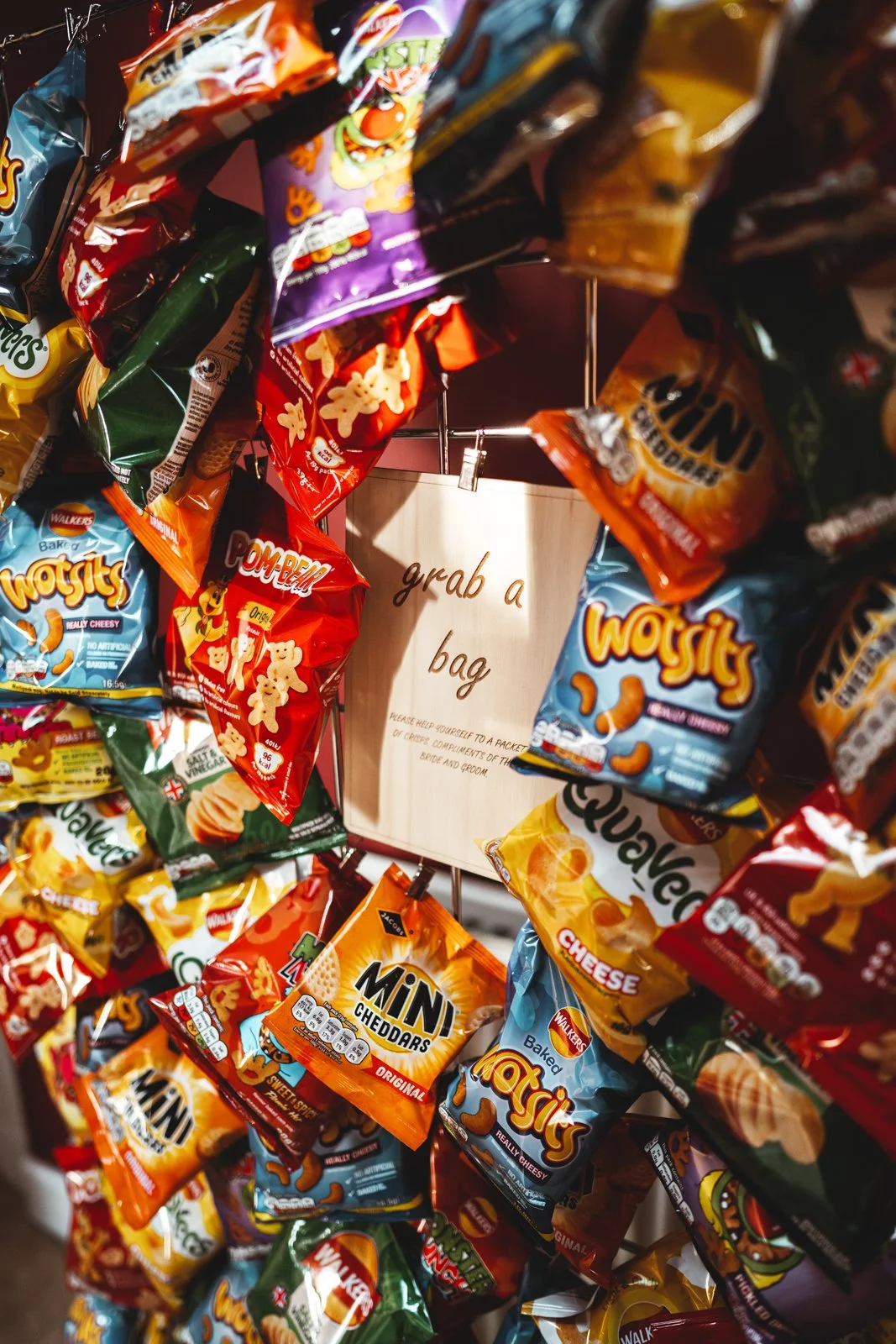 A variety of snack bags, including Wotsits, Pom-Bear, Mini Cheddars, and Walker's crisps, hanging on a display rack with a sign that reads "grab a bag" in the background.