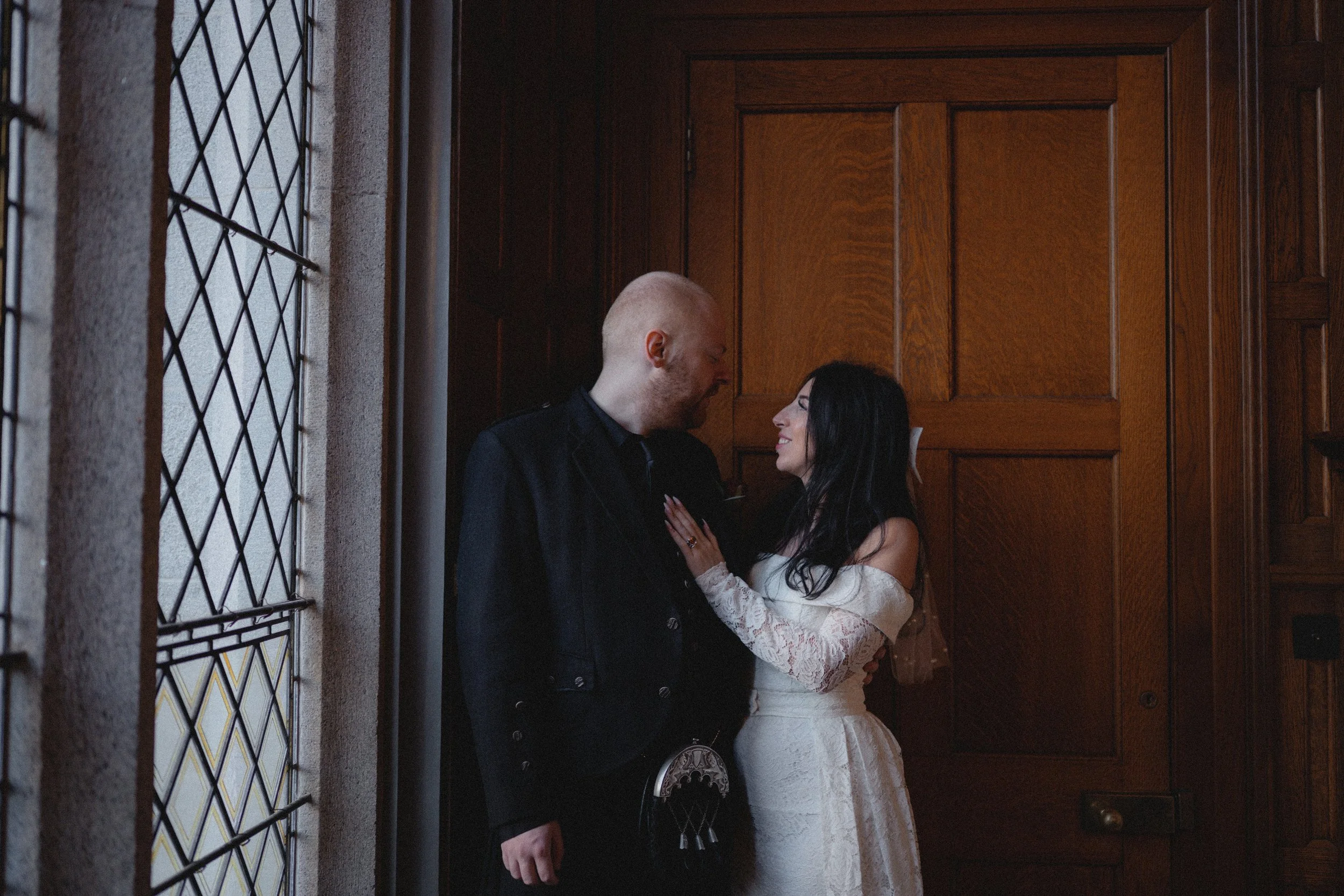A man and woman standing close together indoors, smiling at each other, with a large wooden door behind them and a stained glass window to the side.