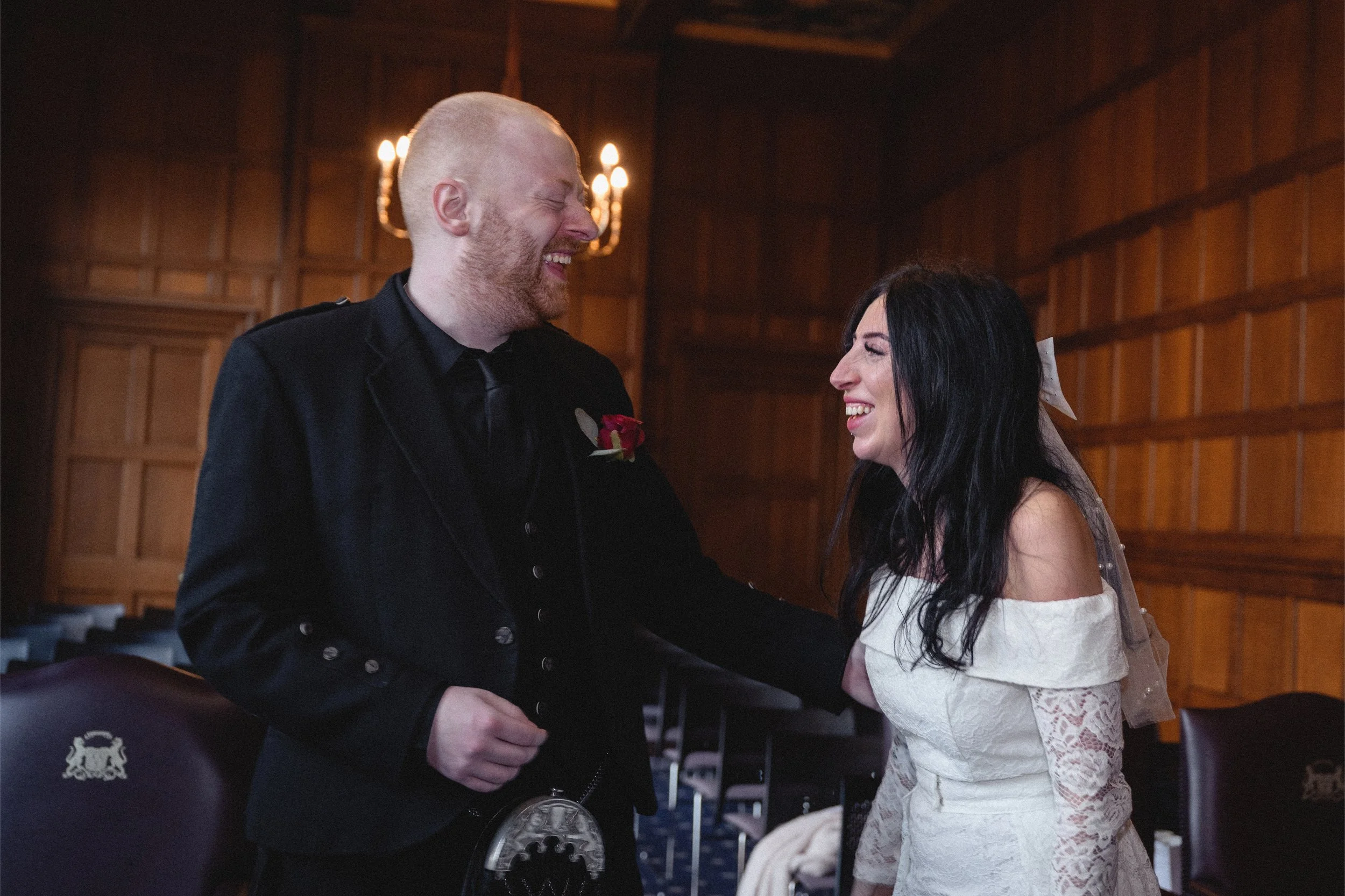 A man and woman are laughing and holding hands in a wood-paneled room, dressed in wedding attire.