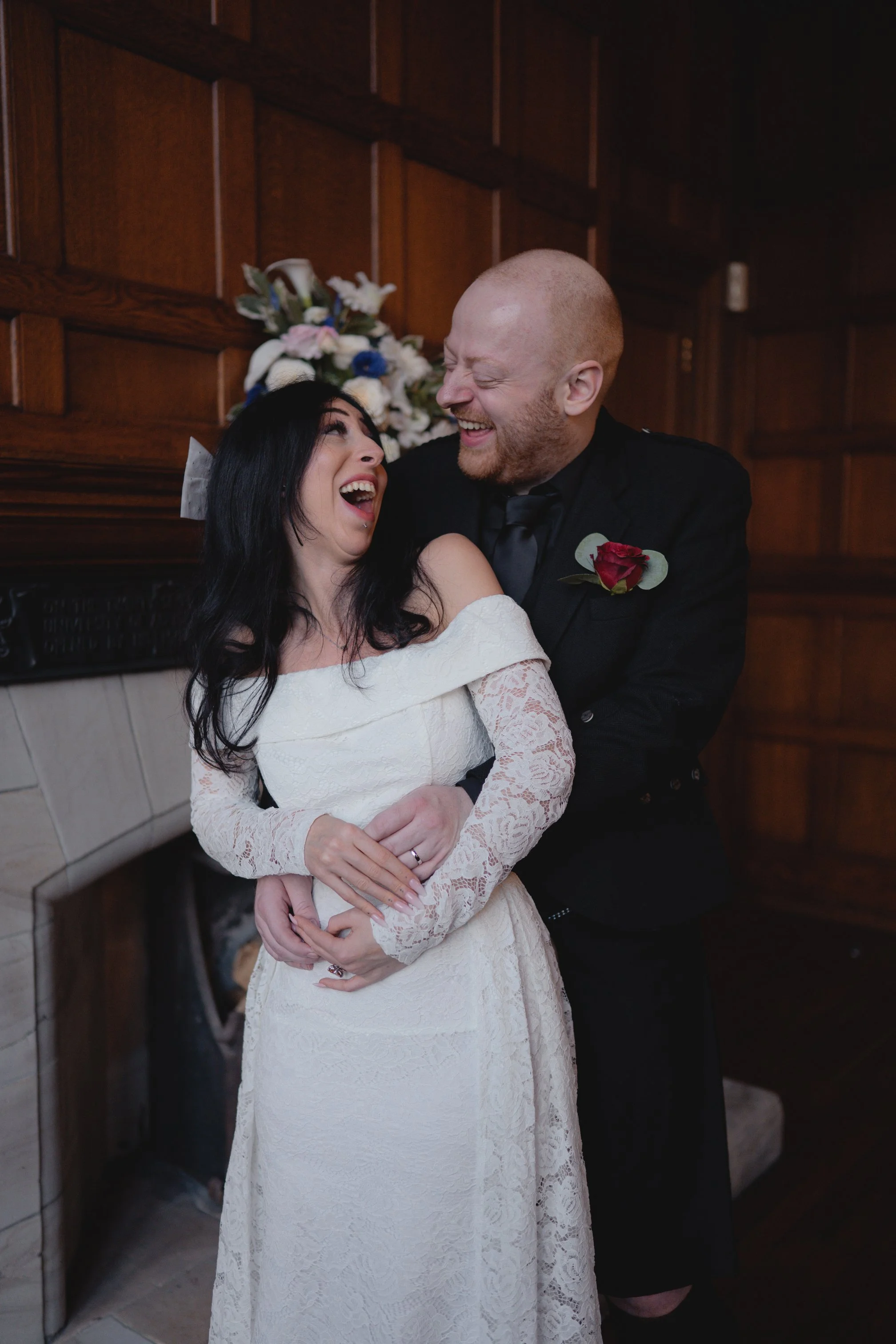A bride and groom sharing a joyful moment during their wedding, standing in front of a fireplace with a floral arrangement in the background.