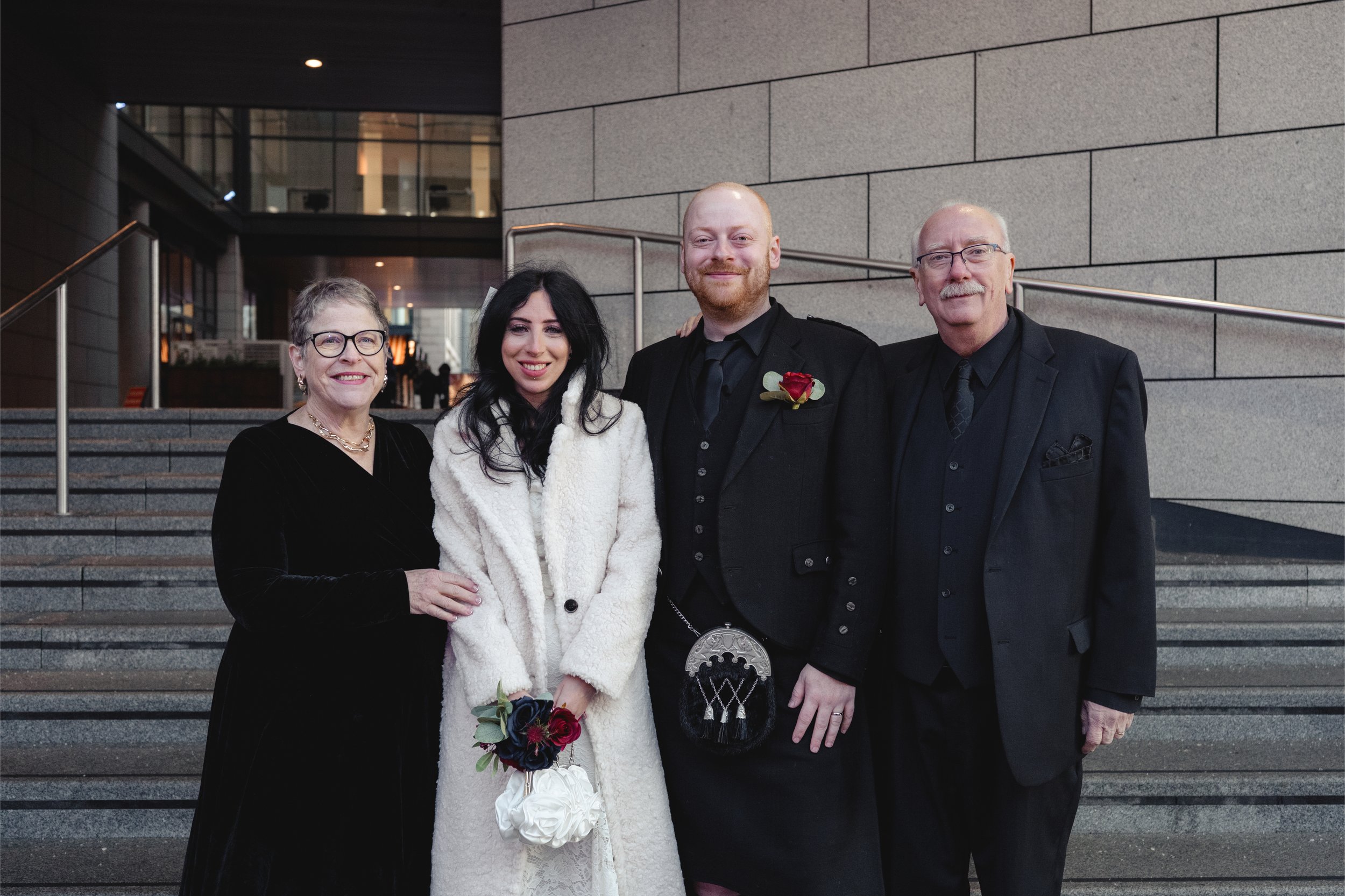 A group of four people dressed in formal attire, standing on steps outside a modern building with large glass windows and a stone wall in the background, at a wedding or formal event.