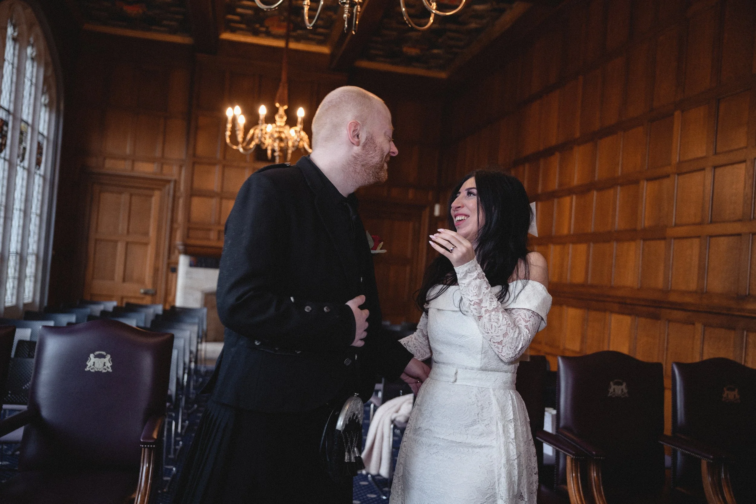 A woman in a white lace dress and a man in a black jacket sharing a joyful moment in a wood-paneled room with chandeliers.
