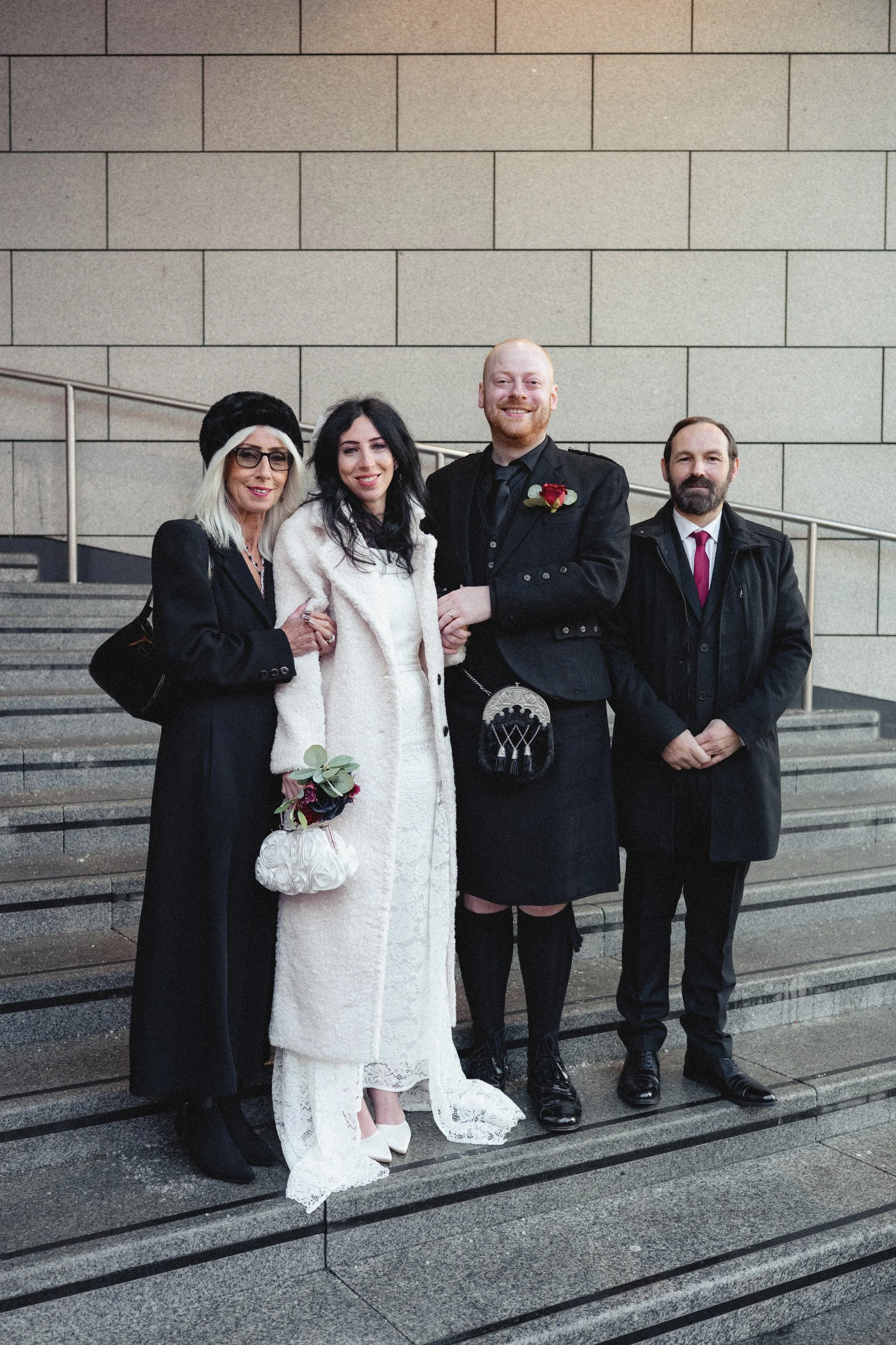 A group of four people standing on stairs in front of a beige wall, dressed in formal and traditional attire, possibly for a wedding or special occasion, with two women holding bouquets.