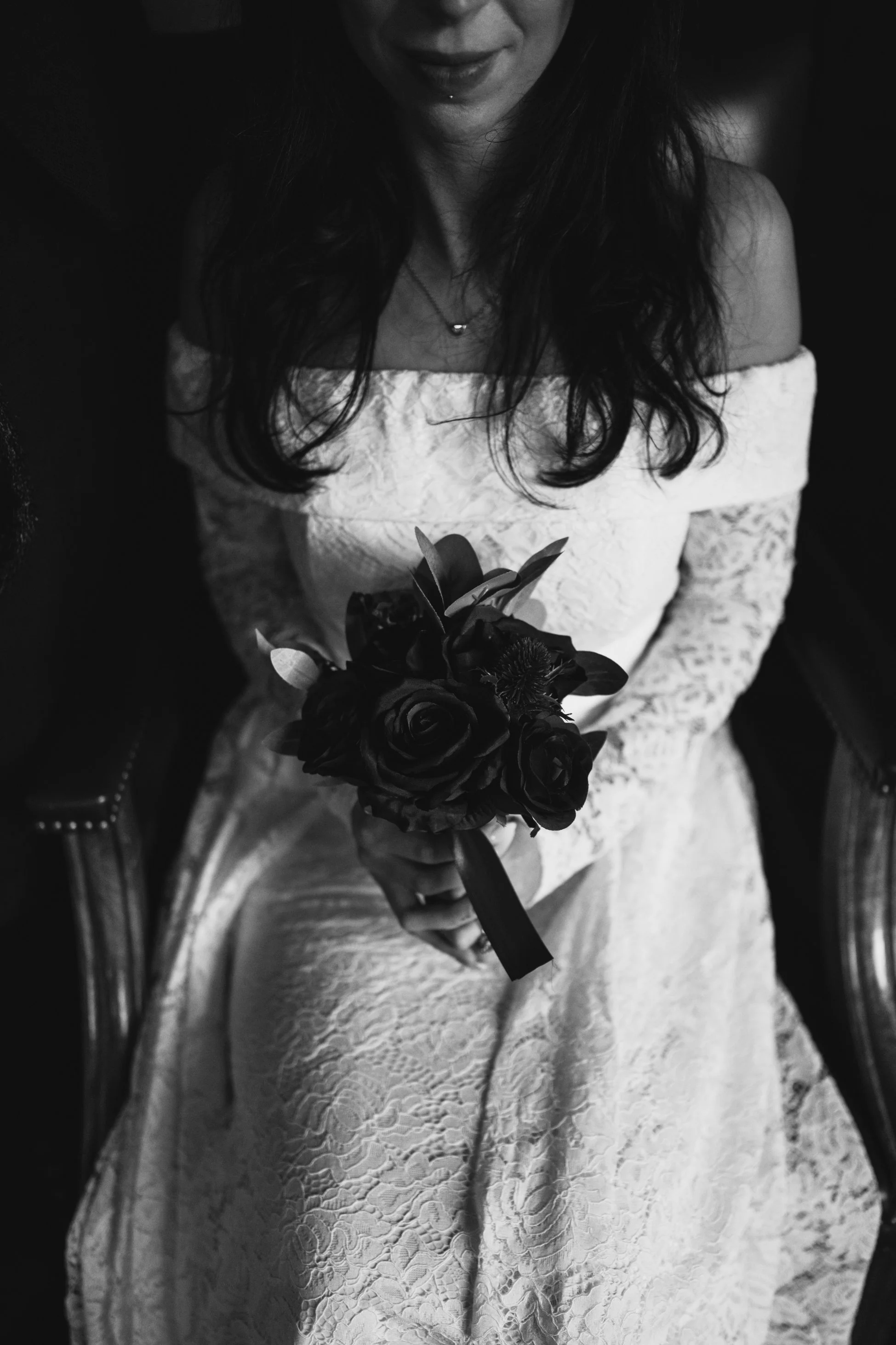A woman in a white lace off-shoulder dress is seated and holding a bouquet of dark roses with greenery, captured in black and white.