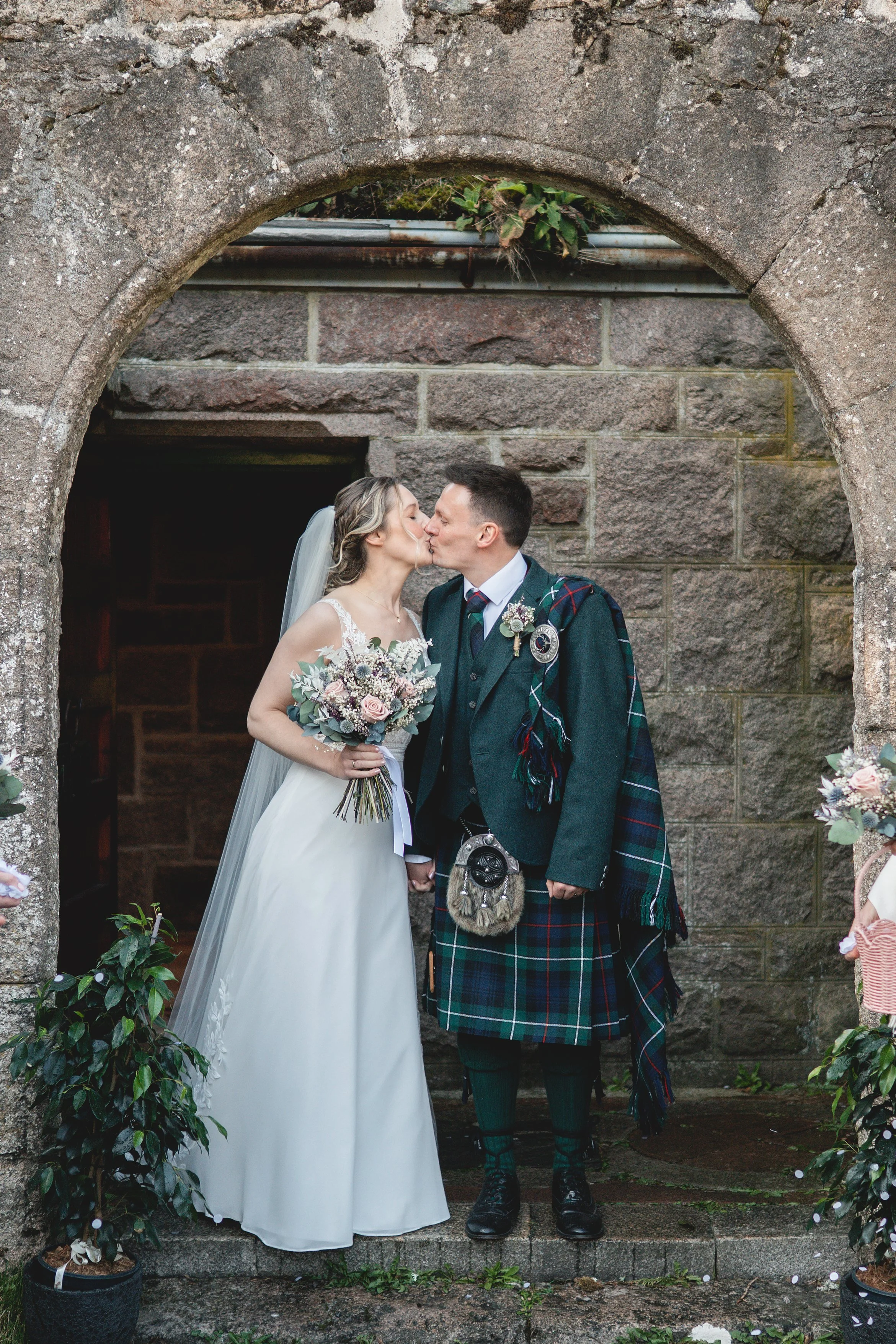 A bride and groom kiss under a stone archway during a wedding ceremony, with greenery and flowers around them.