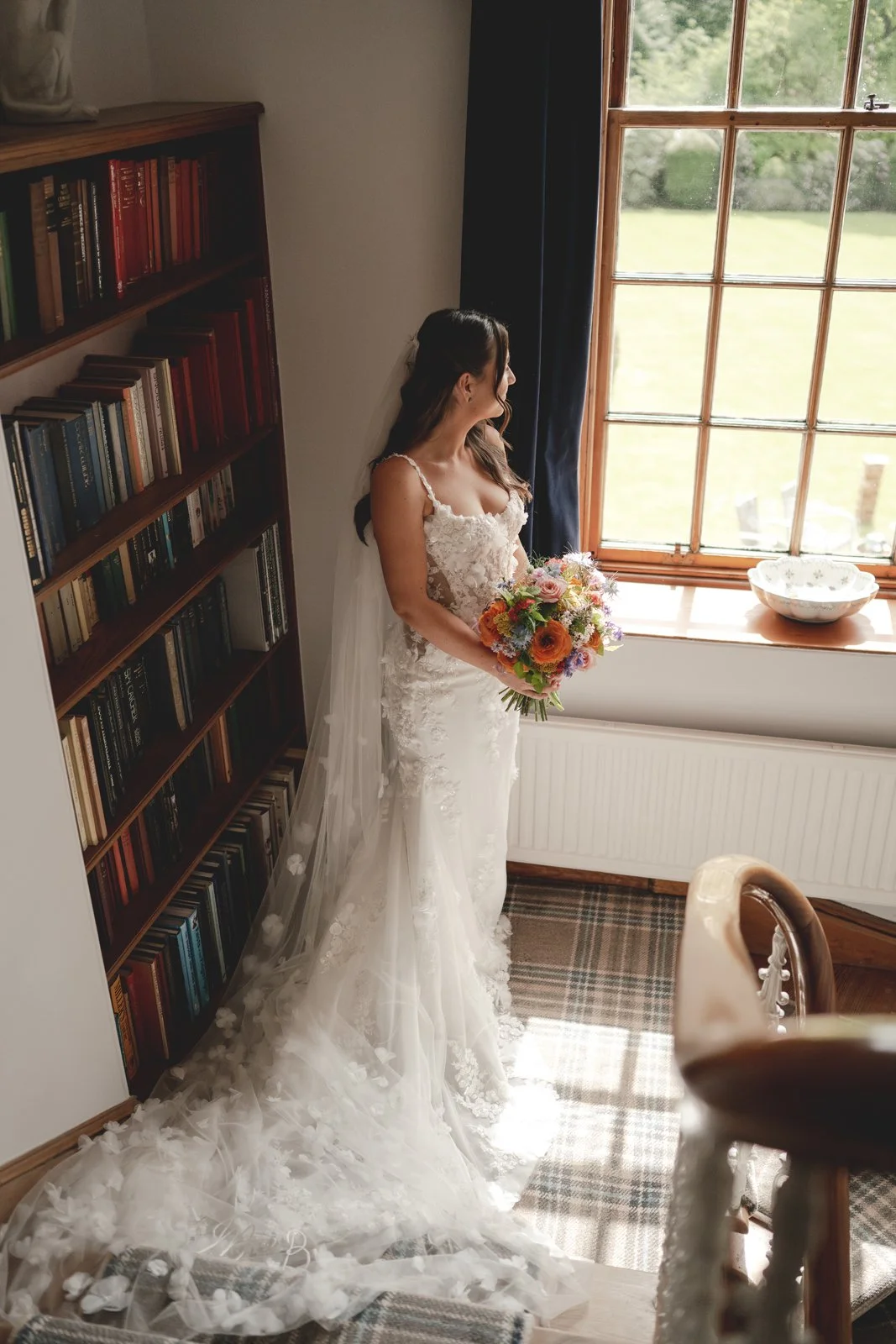 A bride in a lace wedding dress holding a bouquet, standing indoors near a window, with a bookshelf and a window with a view of a garden outside.