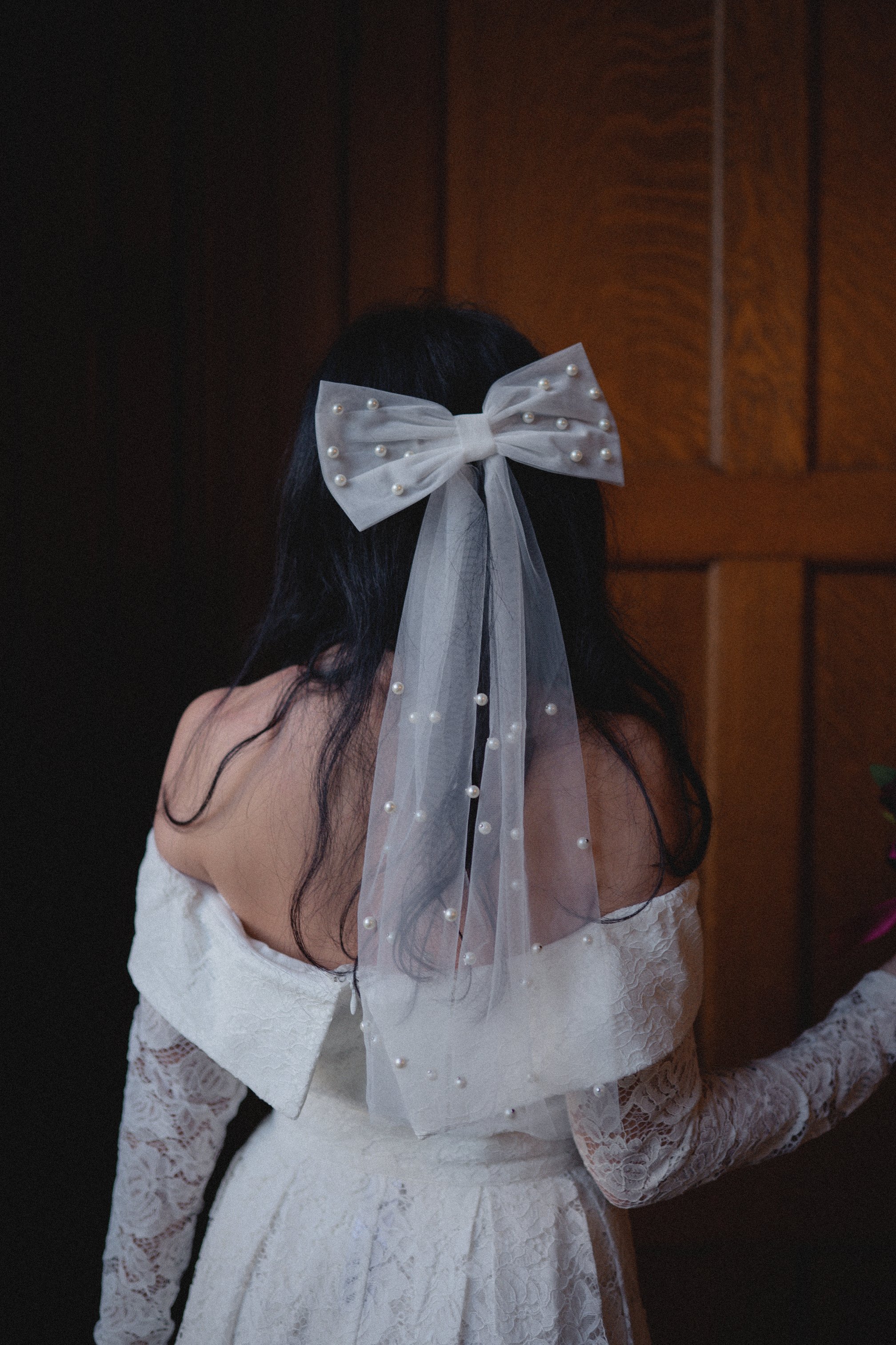A woman in a wedding dress has a large white bow with pearl embellishments in her dark hair, standing near a wooden wall.