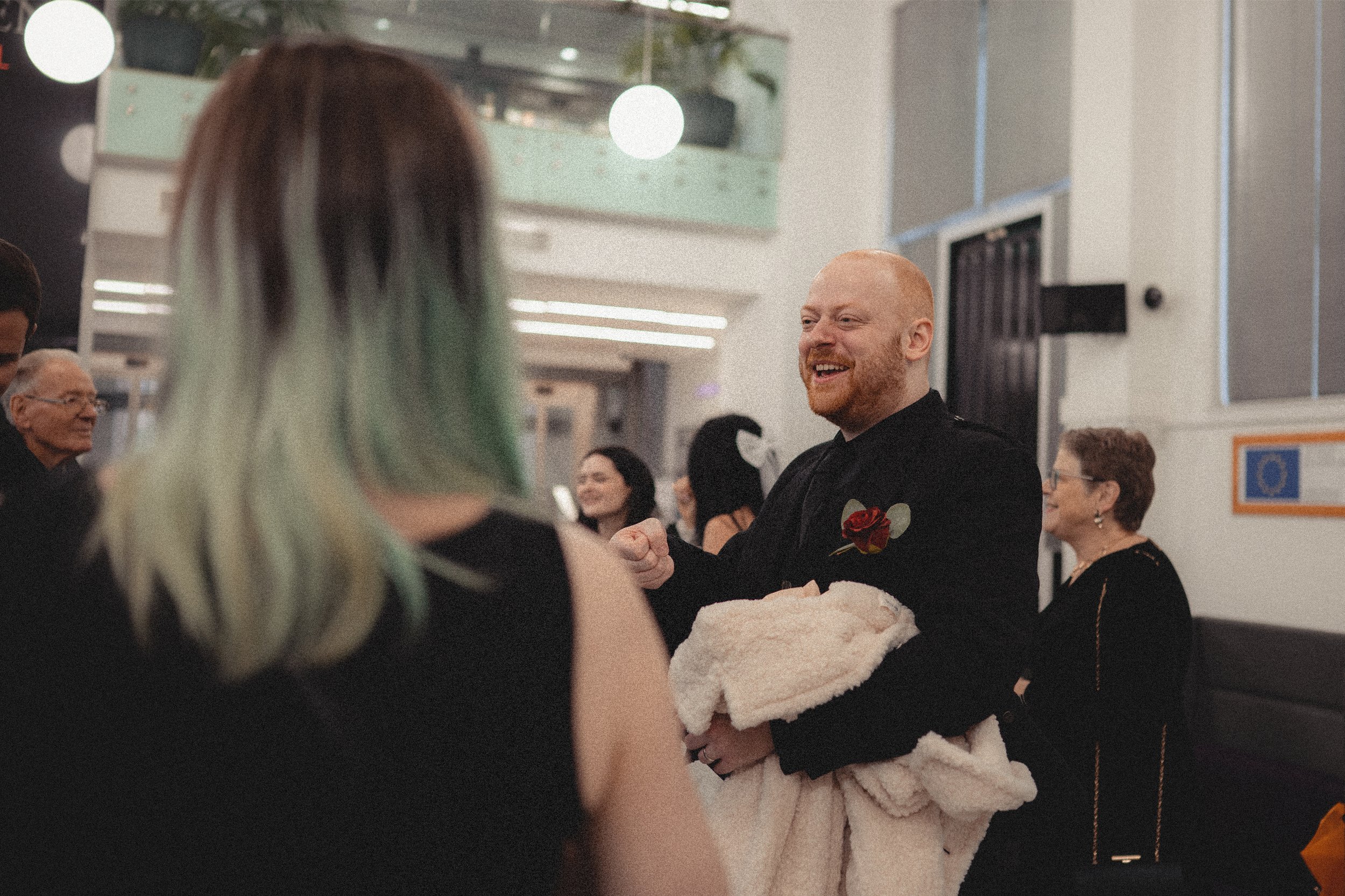 A man with a red beard and a boutonniere on his chest is smiling and talking to a woman with multi-colored hair at an indoor event. The man is holding a fluffy blanket or towel.