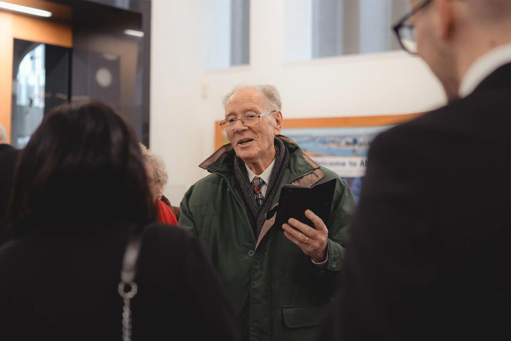 An elderly man speaking to a group of people indoors, holding a tablet and wearing glasses, a green jacket, and a patterned tie.