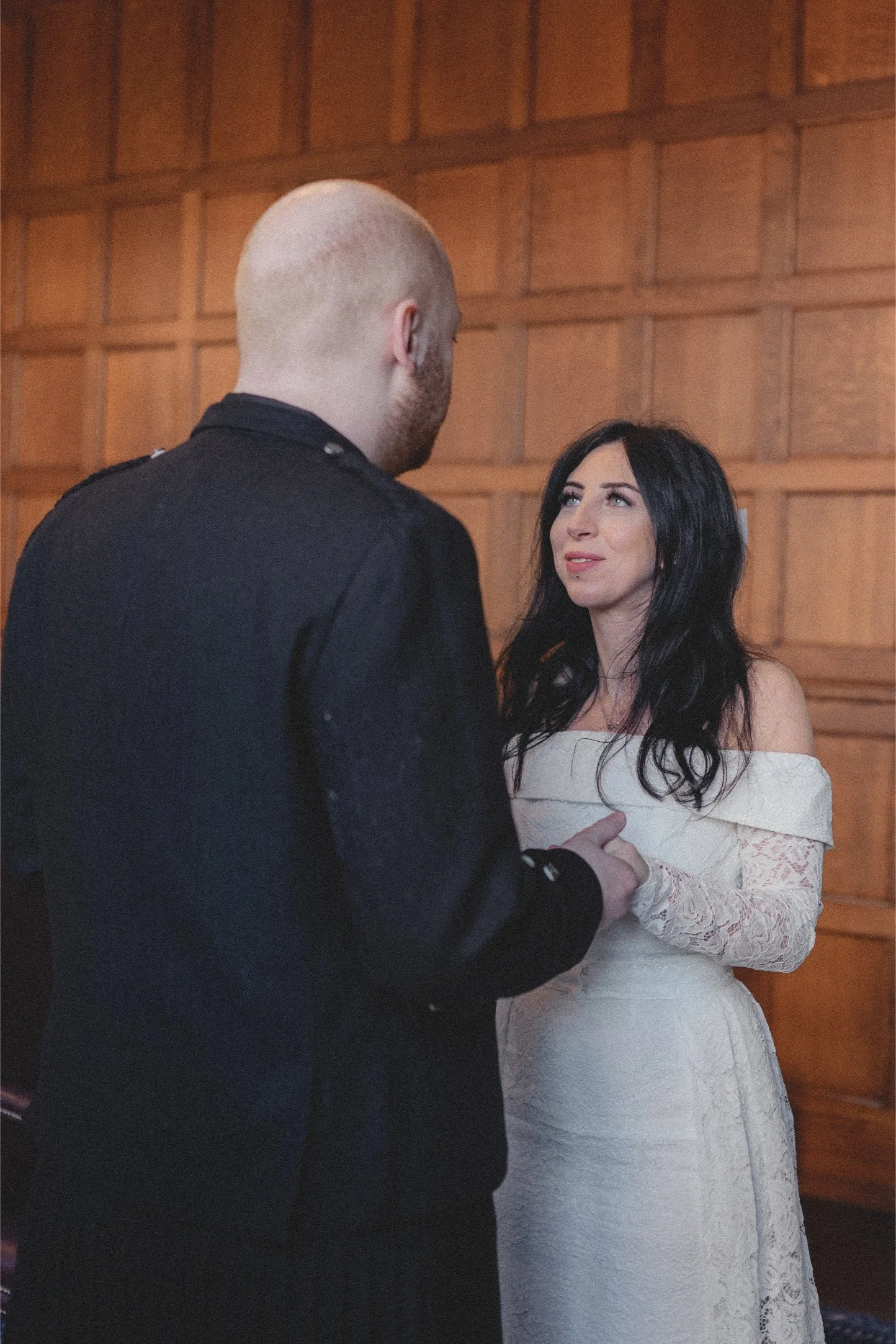 A woman with black hair wearing a white off-shoulder lace dress looks at a man with a shaved head dressed in black, holding her hand, in a wooden-paneled room.